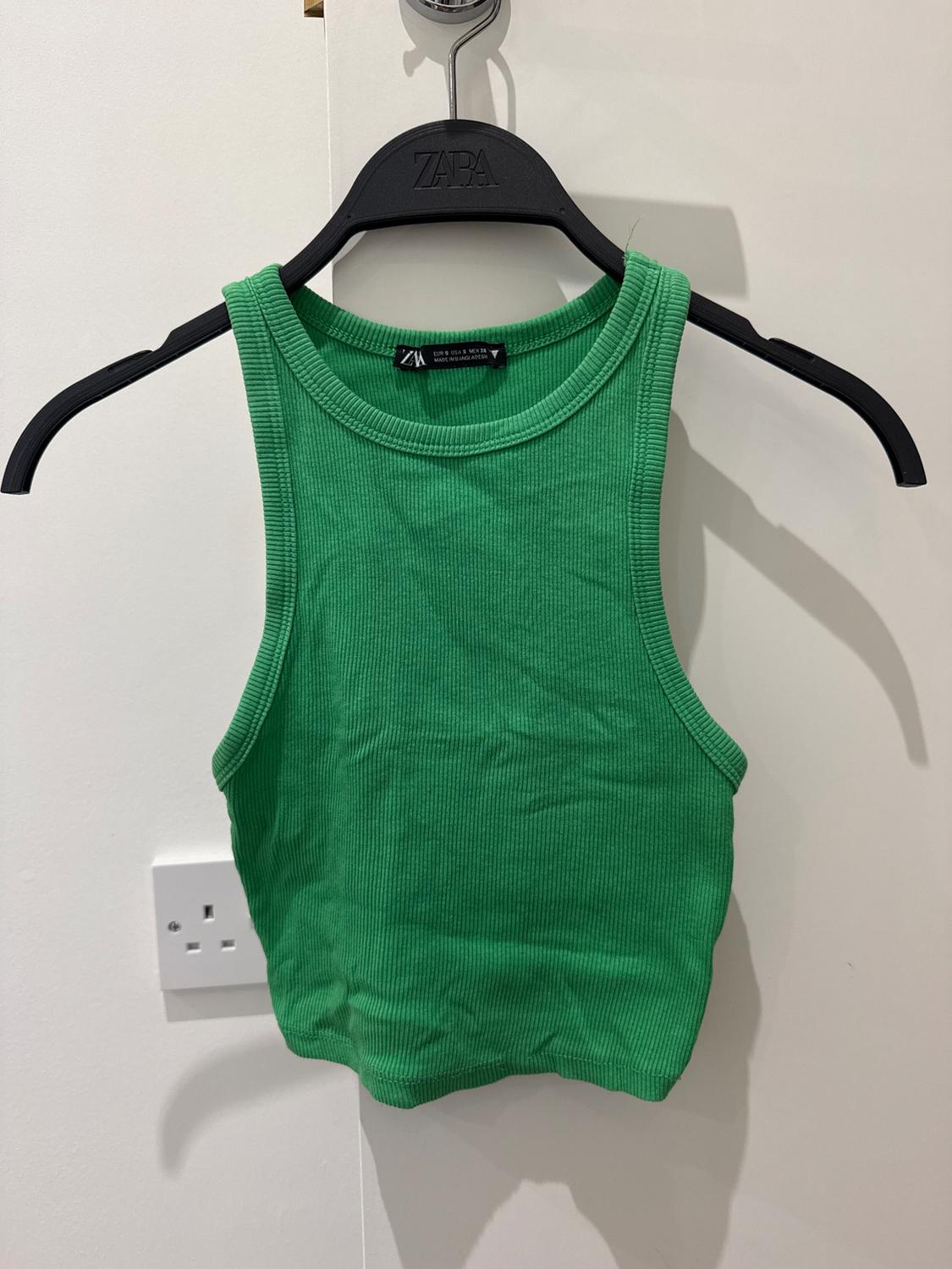 Green sleeveless top hanging on a black hanger with the brand visible. White background with a plug socket on the wall.