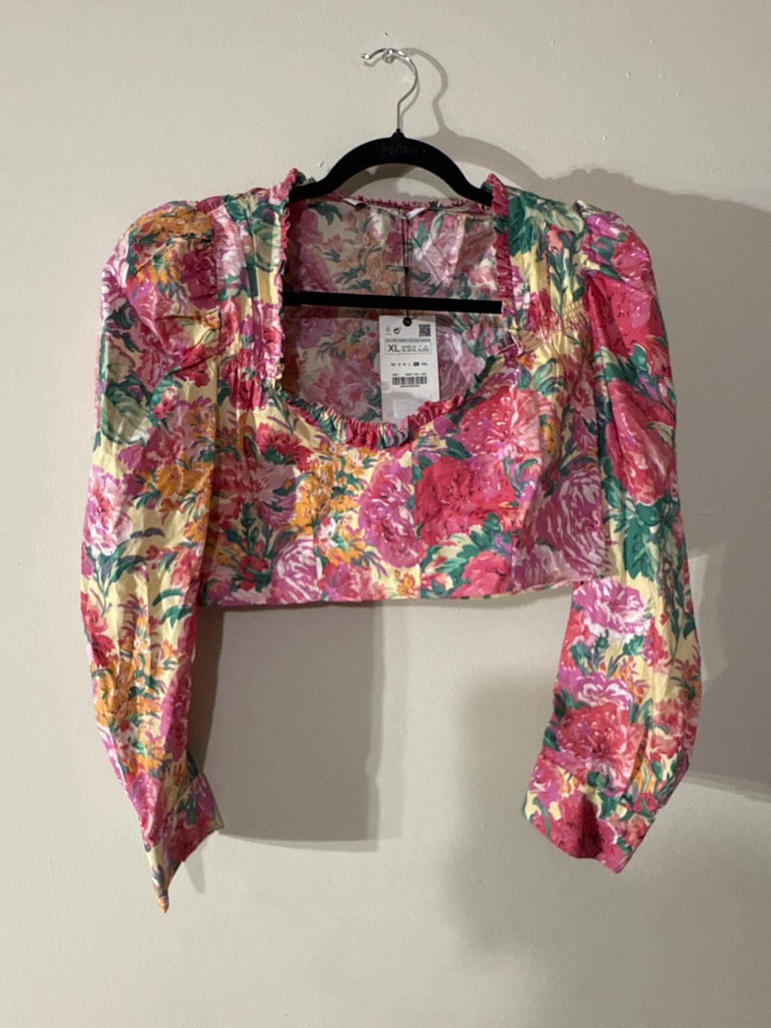 Cropped long-sleeve blouse with a floral print in shades of pink, yellow, and green, hanging on a black hanger against a white background.