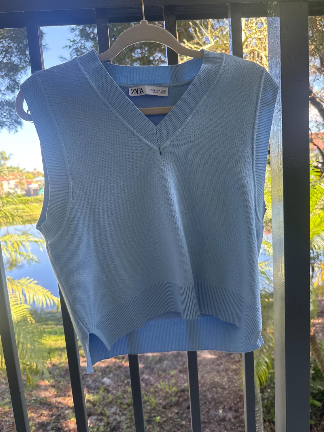 Light blue sleeveless vest hanging on a hanger, with a visible "ZARA" tag, nature background and black railing.