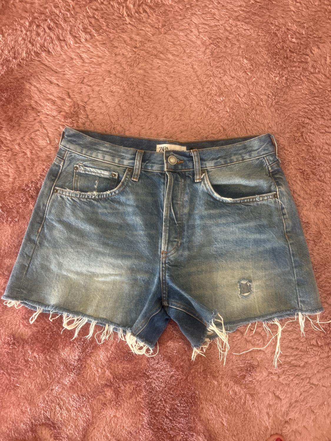 Distressed blue denim shorts with frayed hem and ZARA label, on a pink faux fur background. Casual style.