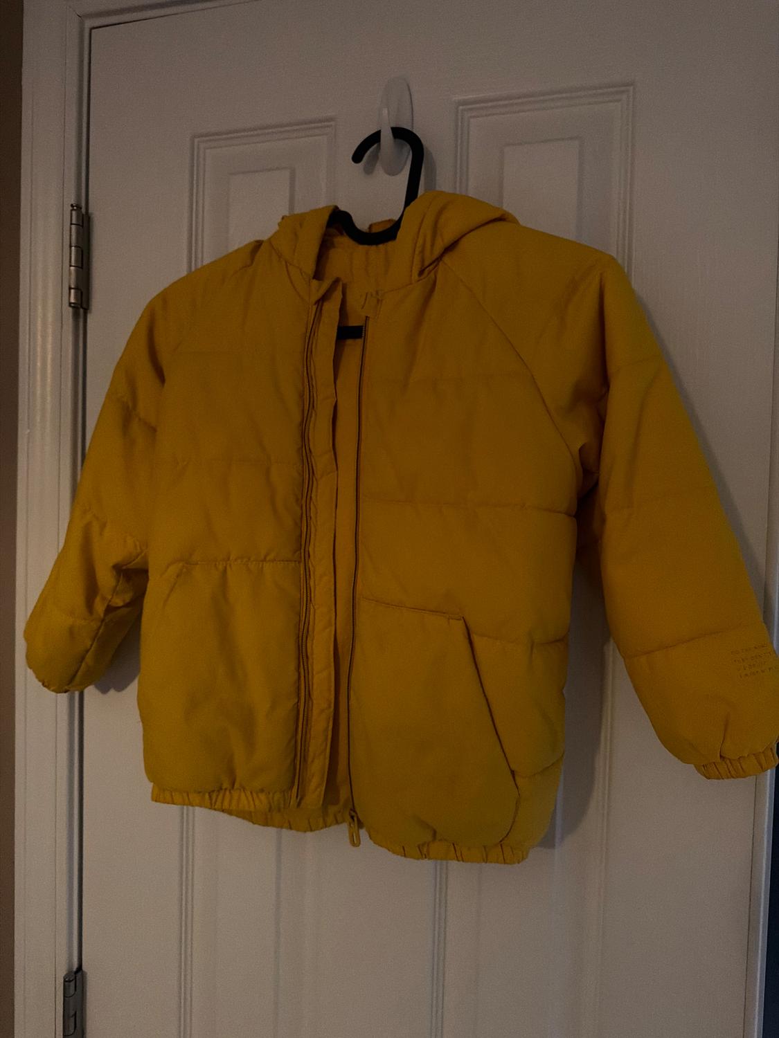 Yellow padded jacket with a hood hanging on a white door. The jacket has pockets and a zipper closure.