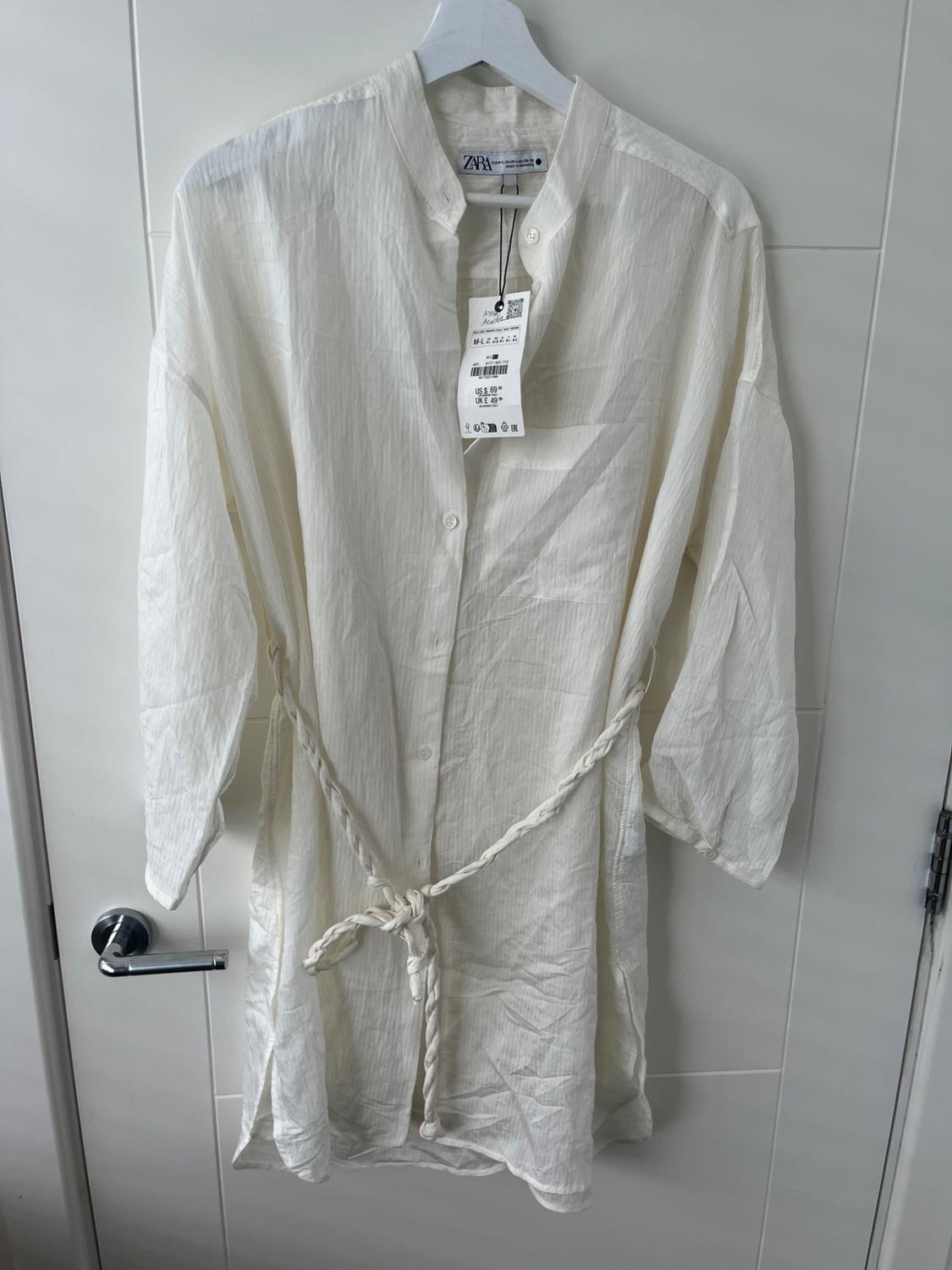 Long-sleeved white shirt with braided belt, hanging on a hanger. Visible label with size and brand details.