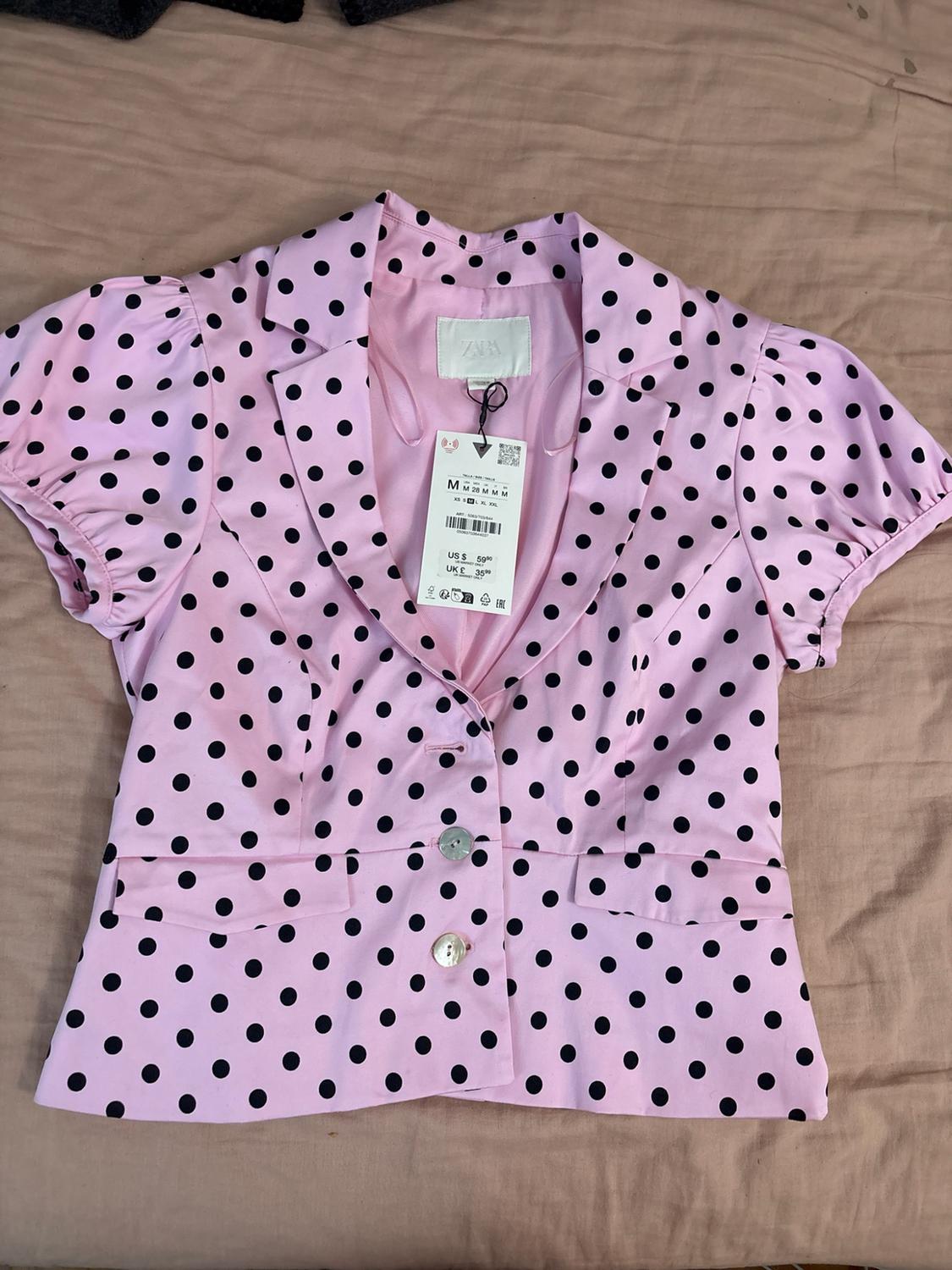 Pink blouse with black polka dots, lapel collar, buttons and short sleeves on beige fabric. Label with ZARA text.