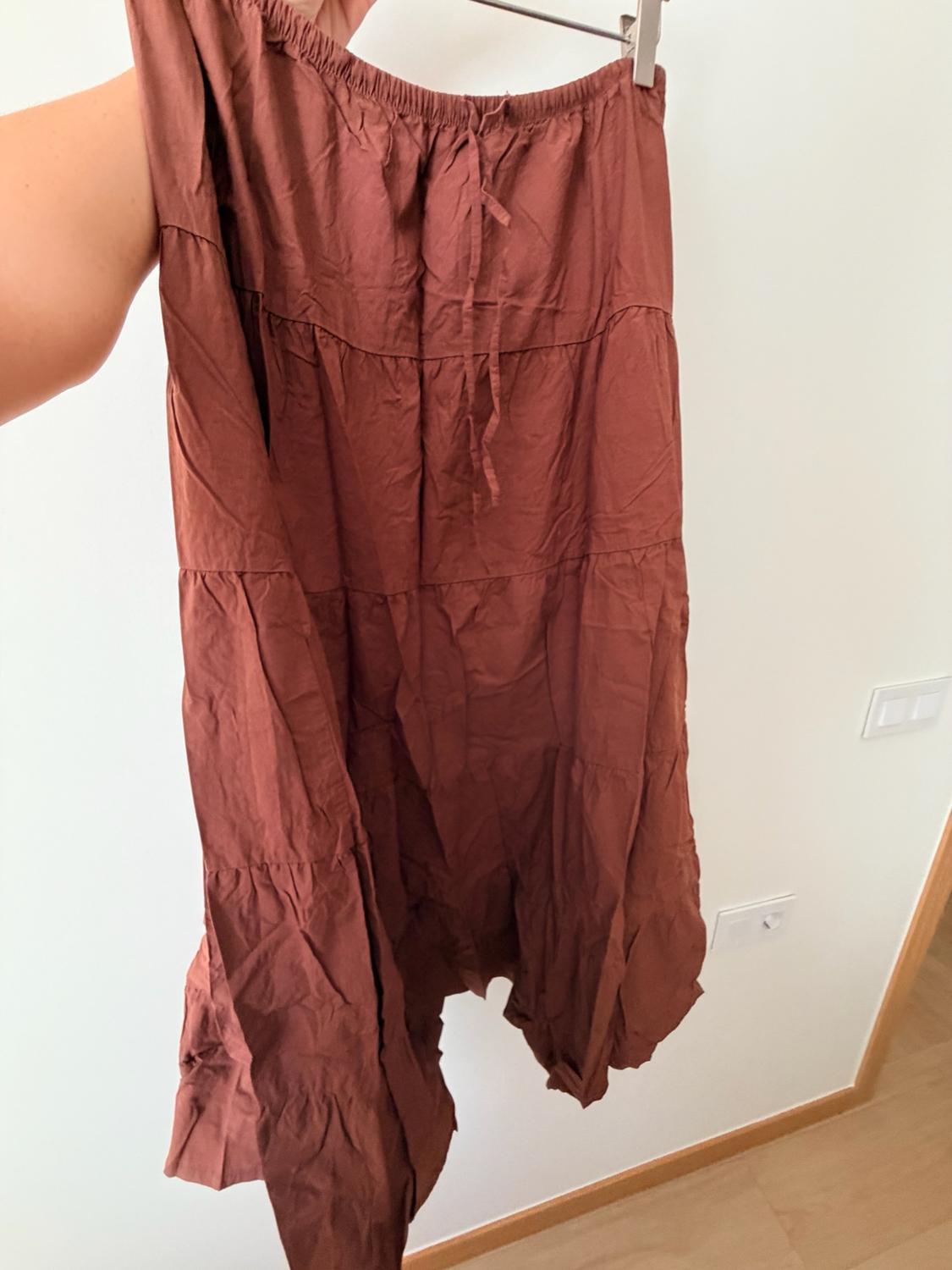 Arm holding a creased brown long ruffled skirt on a hanger in front of a white wall.