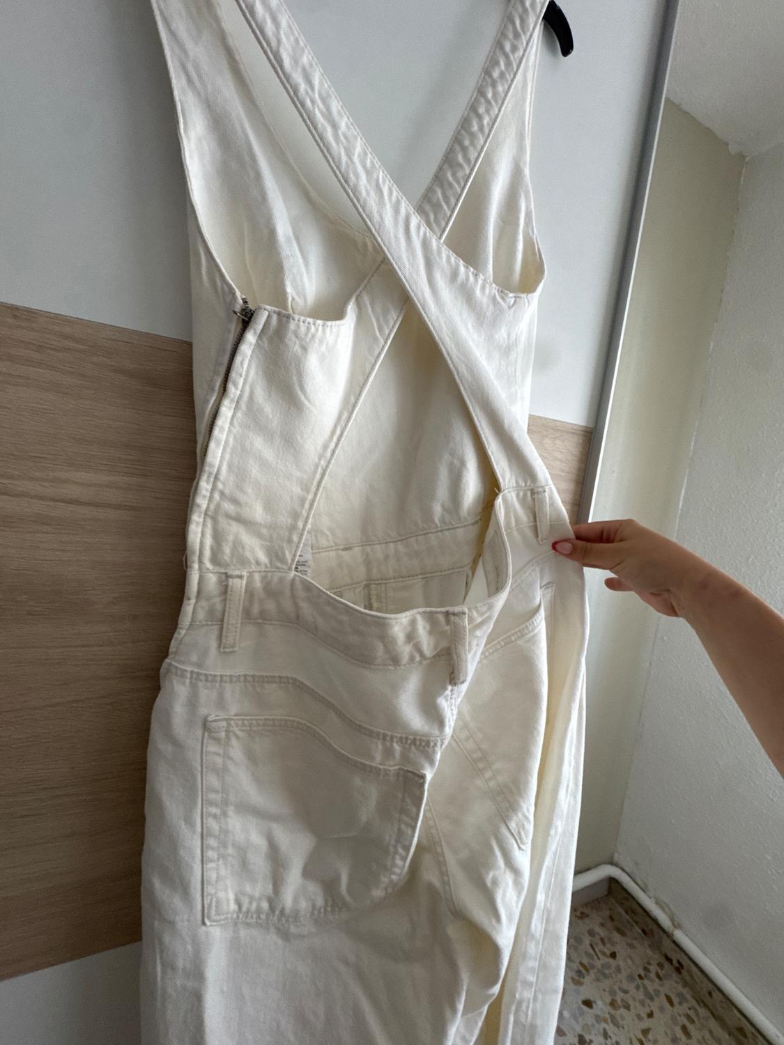 A white jumpsuit with cross-over straps and pockets is hanging on a hanger, held by a hand in an indoor setting.