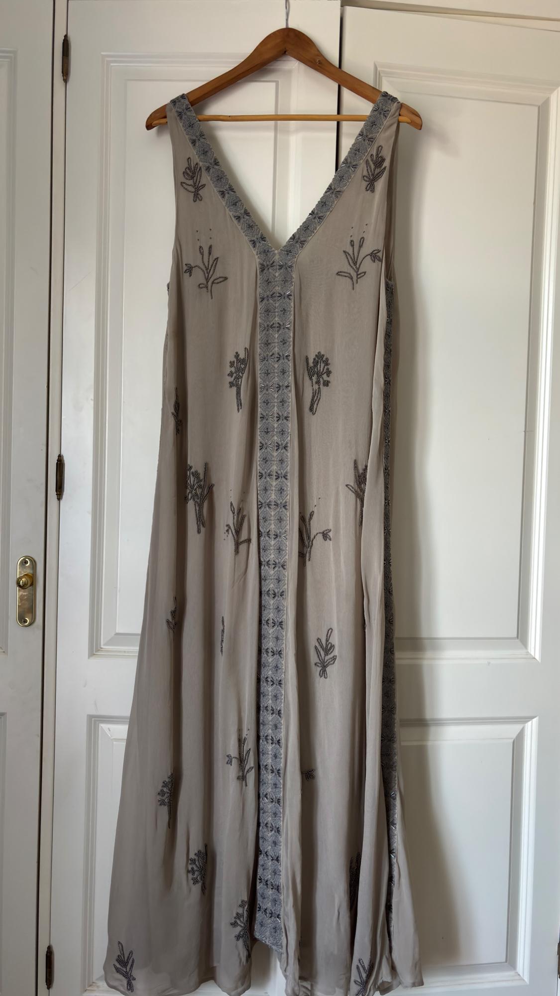 Beige sleeveless maxi dress with a V-neck and floral embroidery, hanging on a wooden hanger over a white door.