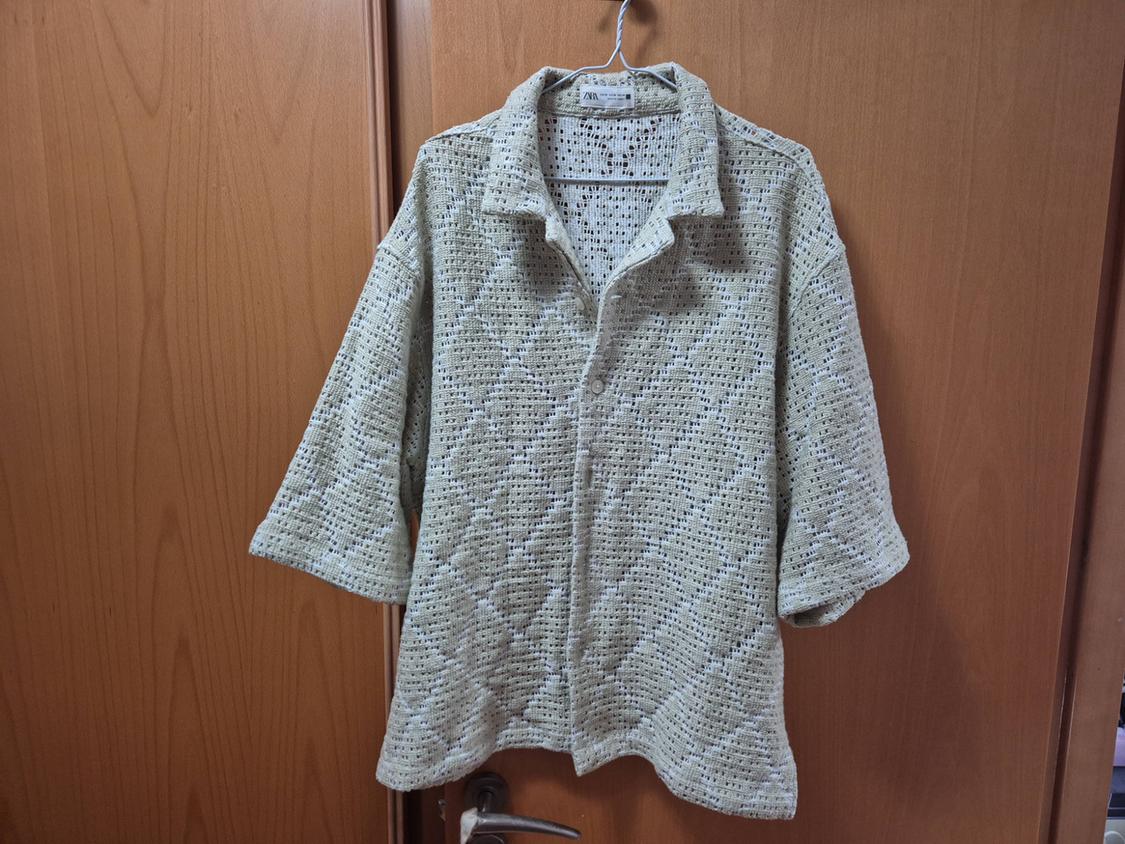 Beige open-knit short-sleeved shirt with diamond pattern and buttons, hanging on a hanger in front of a wooden door. ZARA label.