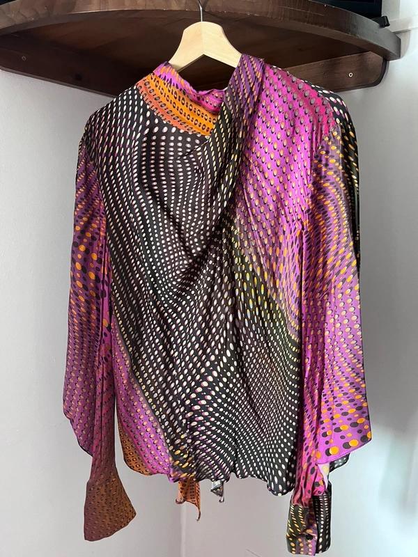 Long-sleeved blouse with a purple, black and orange optical dot print, hanging on a wooden hanger against a white wall.