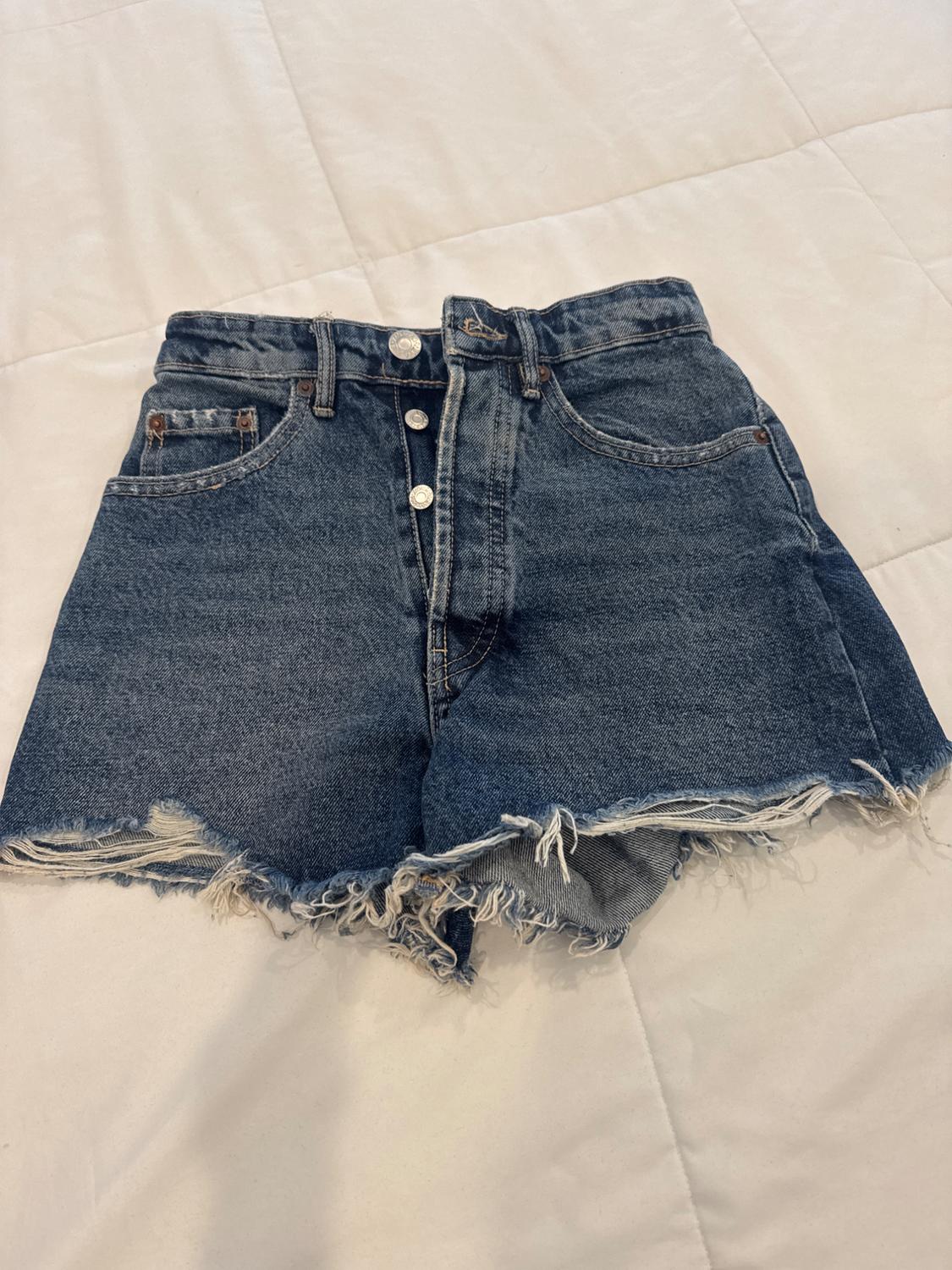 Blue denim shorts with front buttons and frayed edges, placed on a white padded surface.