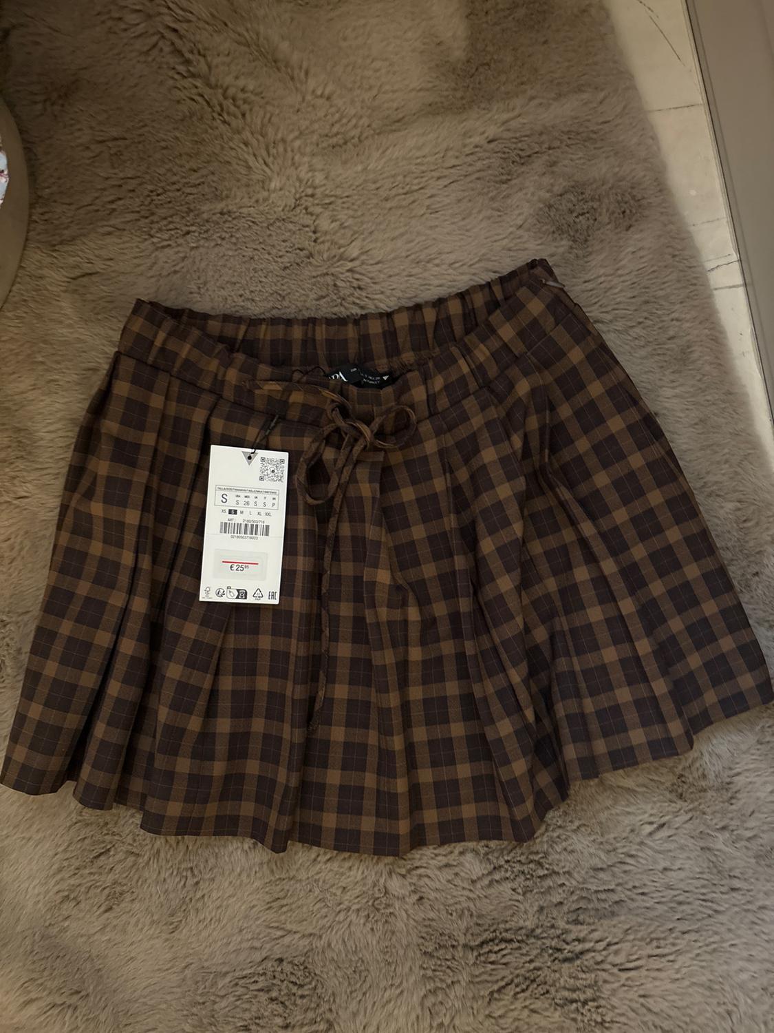 Brown check skirt with a bow at the waist on a beige carpet. Visible tag with a price of €25.