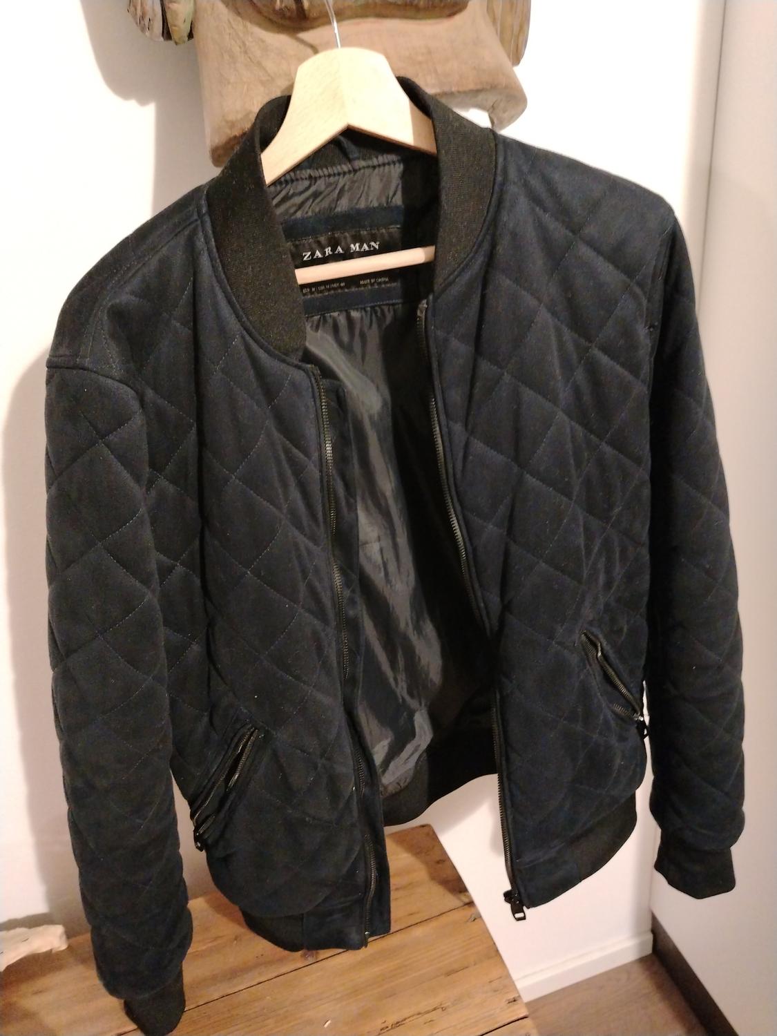 Black quilted jacket hanging on a wooden hanger. Visible inner label with "ZARA MAN" text. White and wood background.