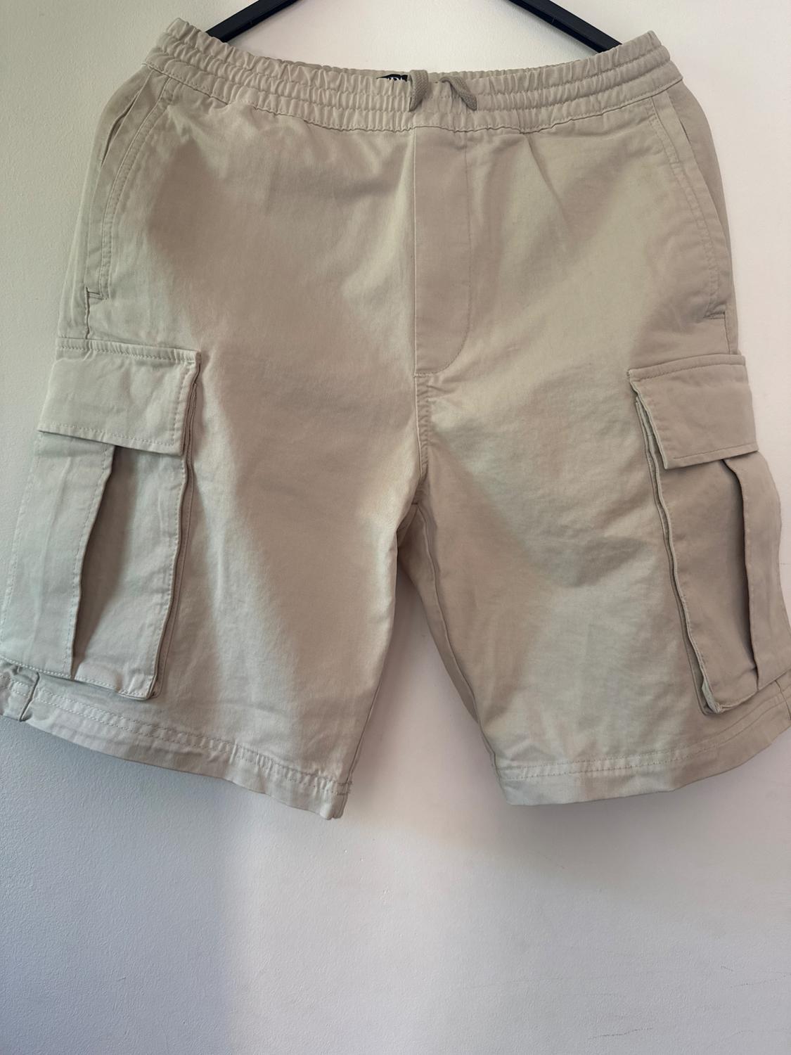Beige cargo shorts with an elasticated waist, drawstring, and side pockets, hanging on a black hanger against a white background.