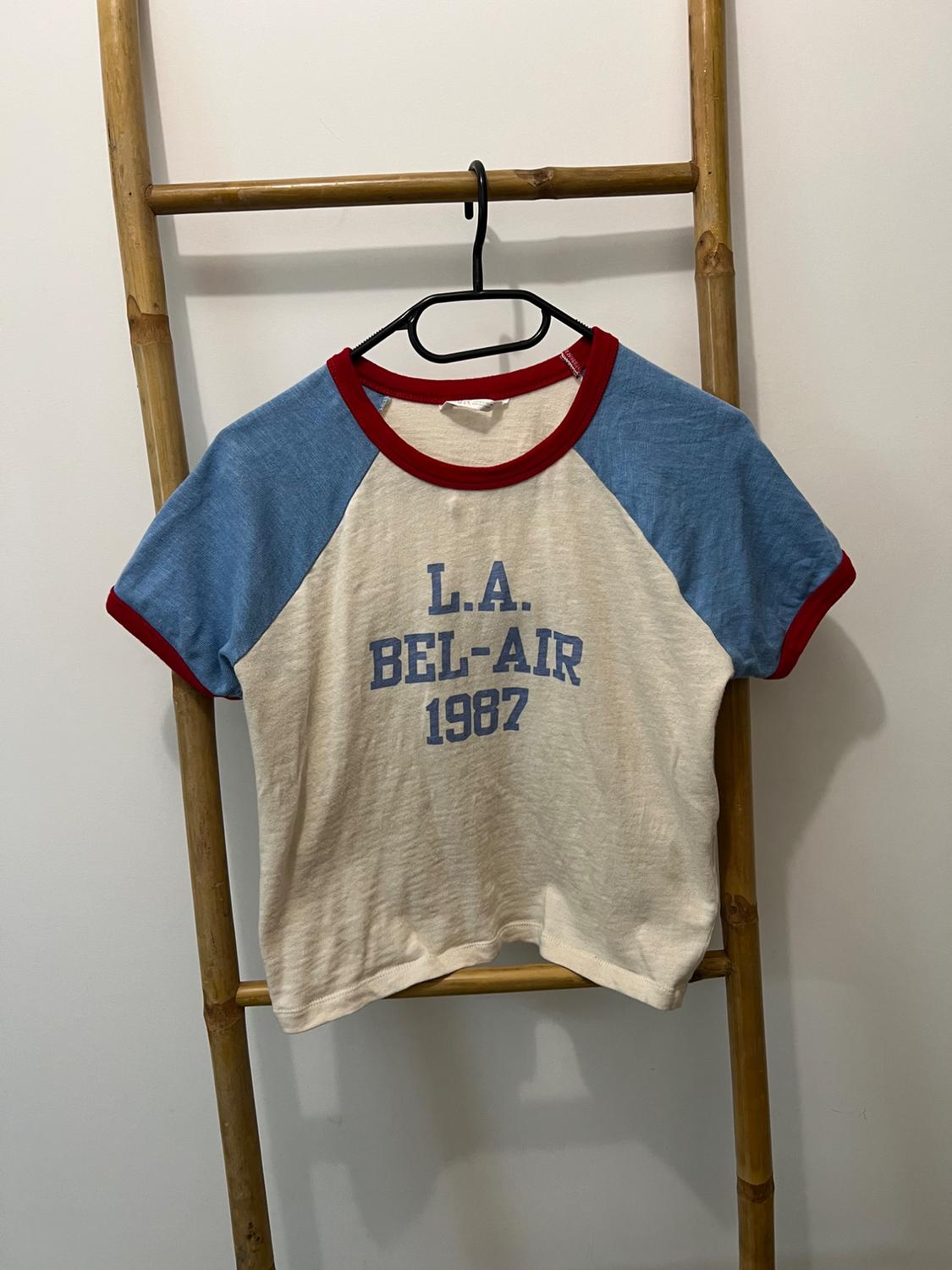 Cream T-shirt with blue sleeves and red trim hanging on a wooden ladder. Blue text L.A. BEL-AIR 1987. Casual style.