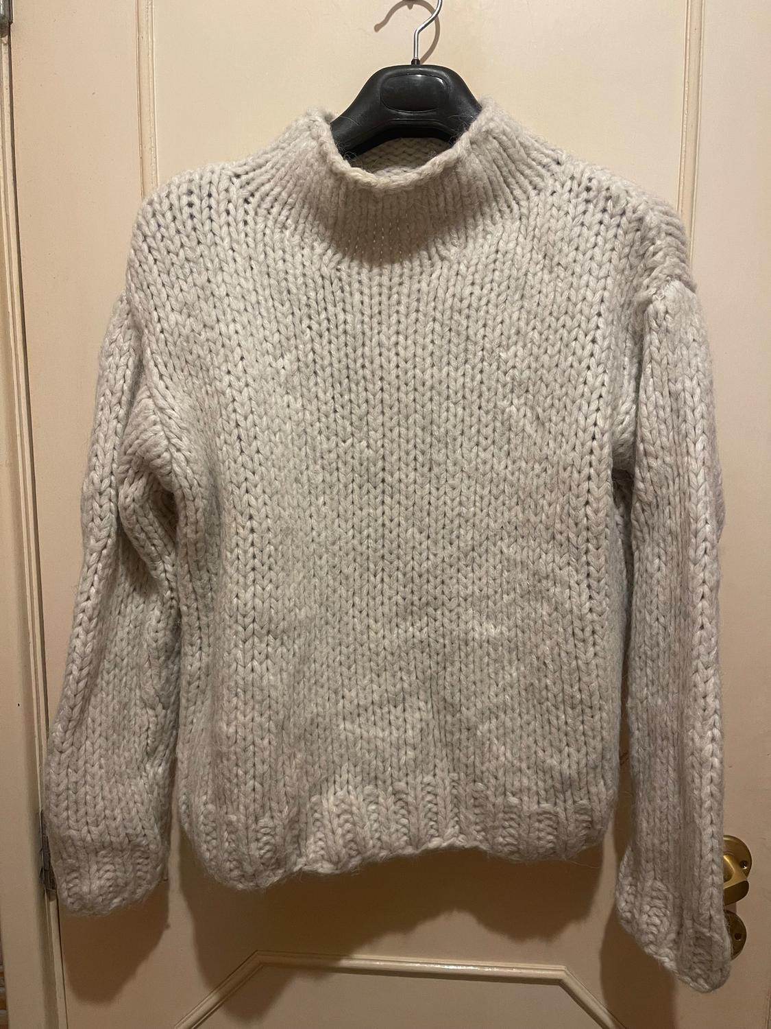 Chunky cream knitted jumper hanging on a black hanger over a beige door.