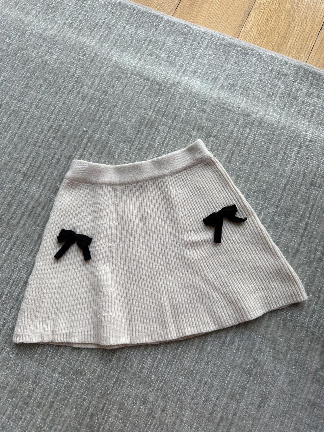 Beige knitted skirt with two black bows, on a grey rug and light wooden floor.