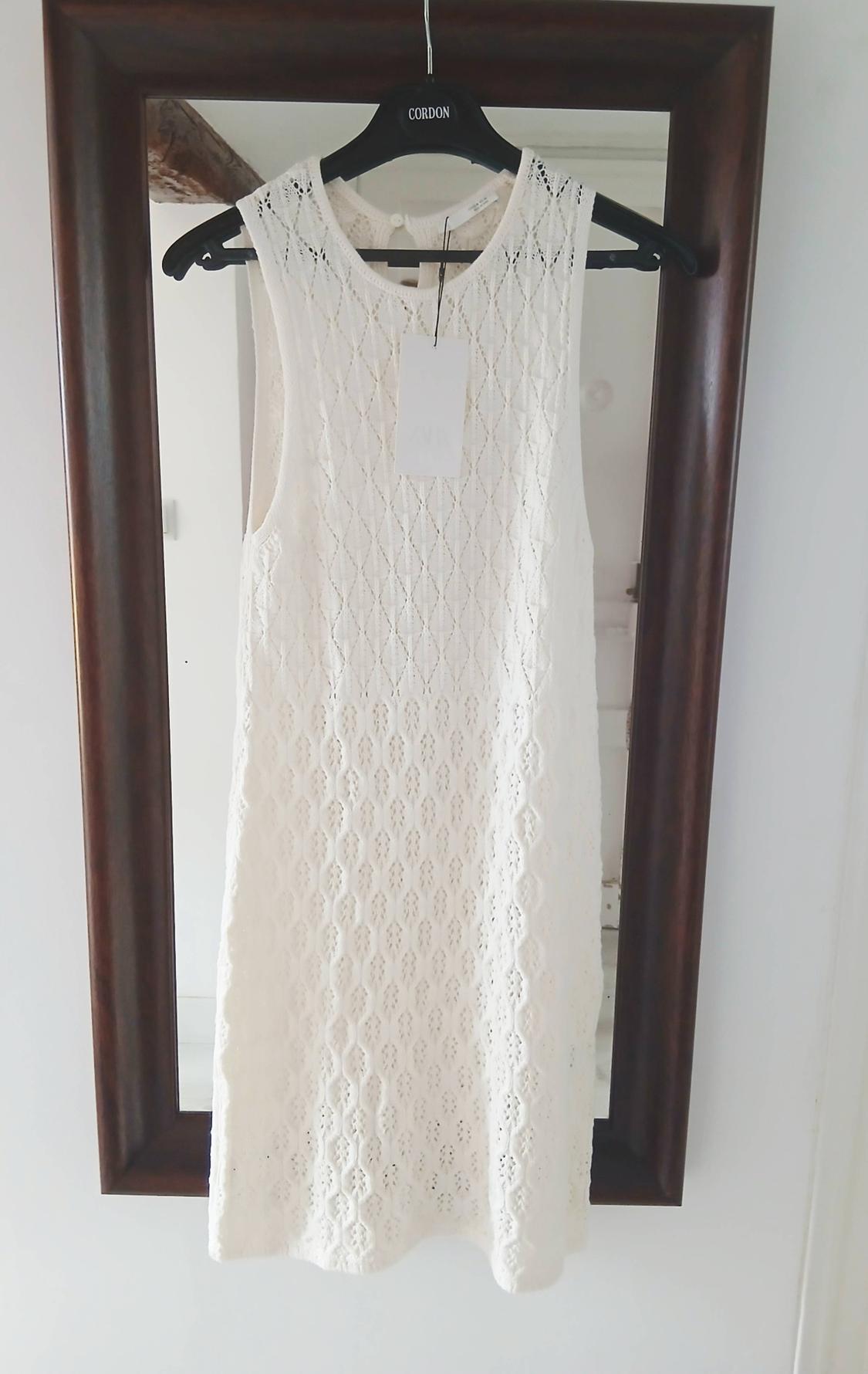 White sleeveless dress with an openwork texture hanging on a black hanger with the text CORDON in front of a wooden-framed mirror.