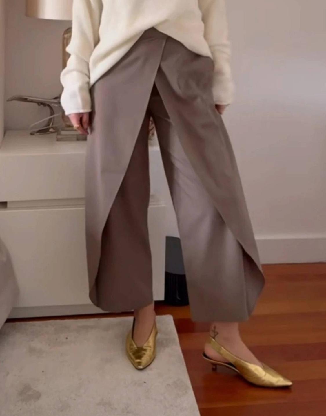 Indoor model wearing a white jumper, taupe trousers with an overlay panel and gold backless shoes. Relaxed setting.