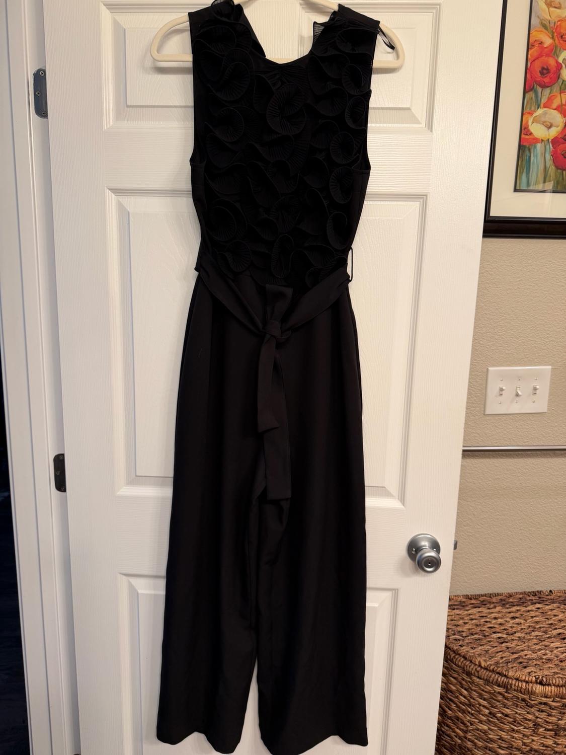 Black sleeveless jumpsuit hanging on a white door. It features a textured circular pattern on the top and a tie at the waist.