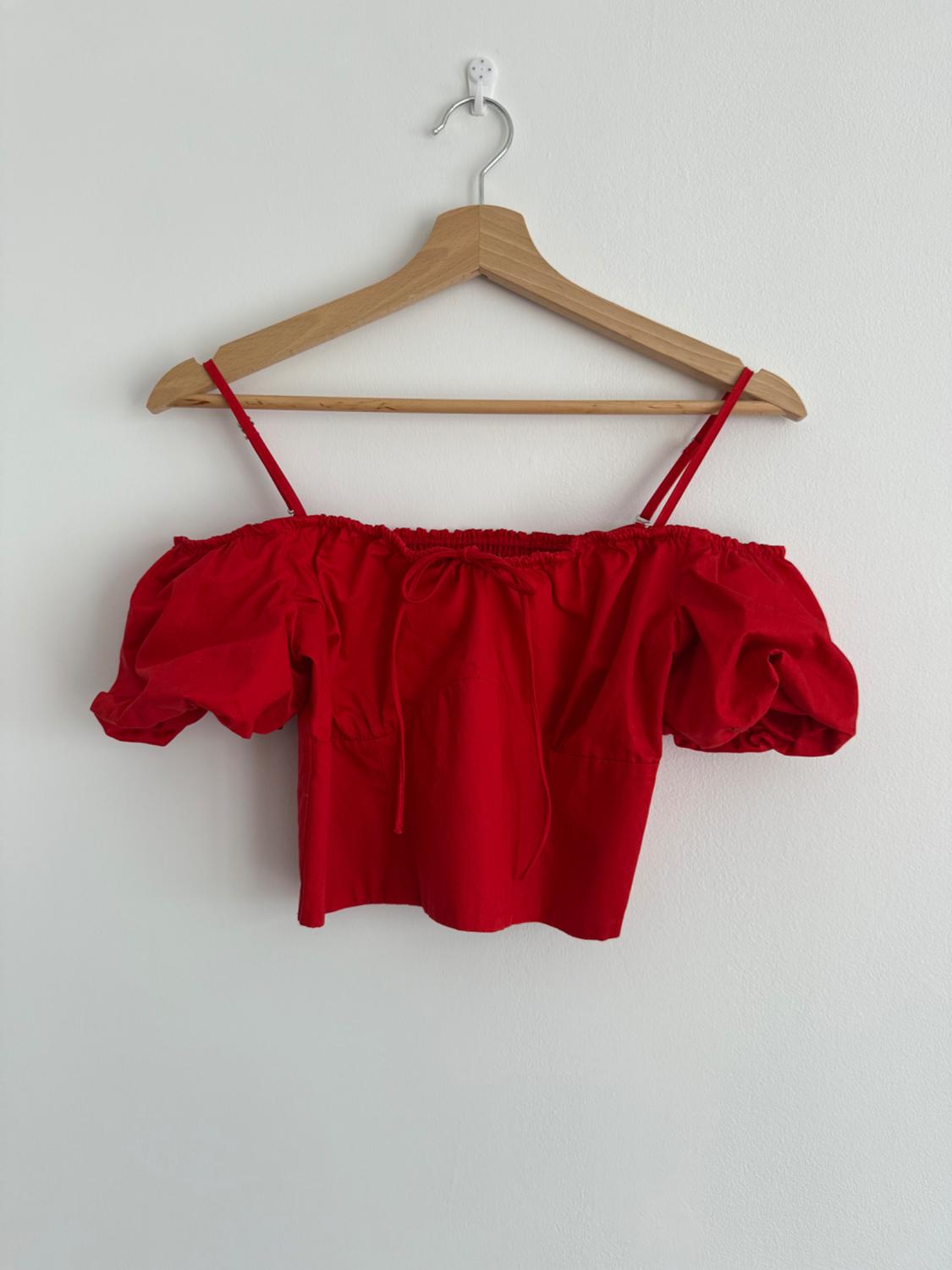 Red blouse with off-the-shoulder neckline and puffed sleeves hanging on a wooden hanger against a white wall.