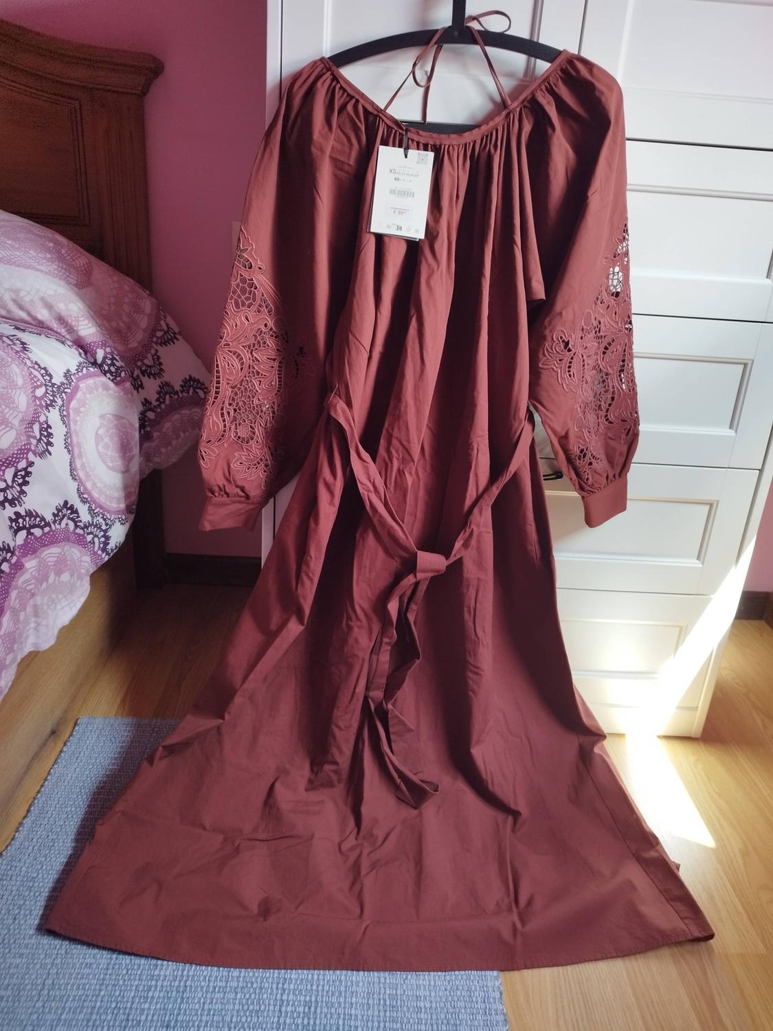 Long brown dress with embroidered sleeves, hanging on a hanger in a room with pink walls and white furniture.