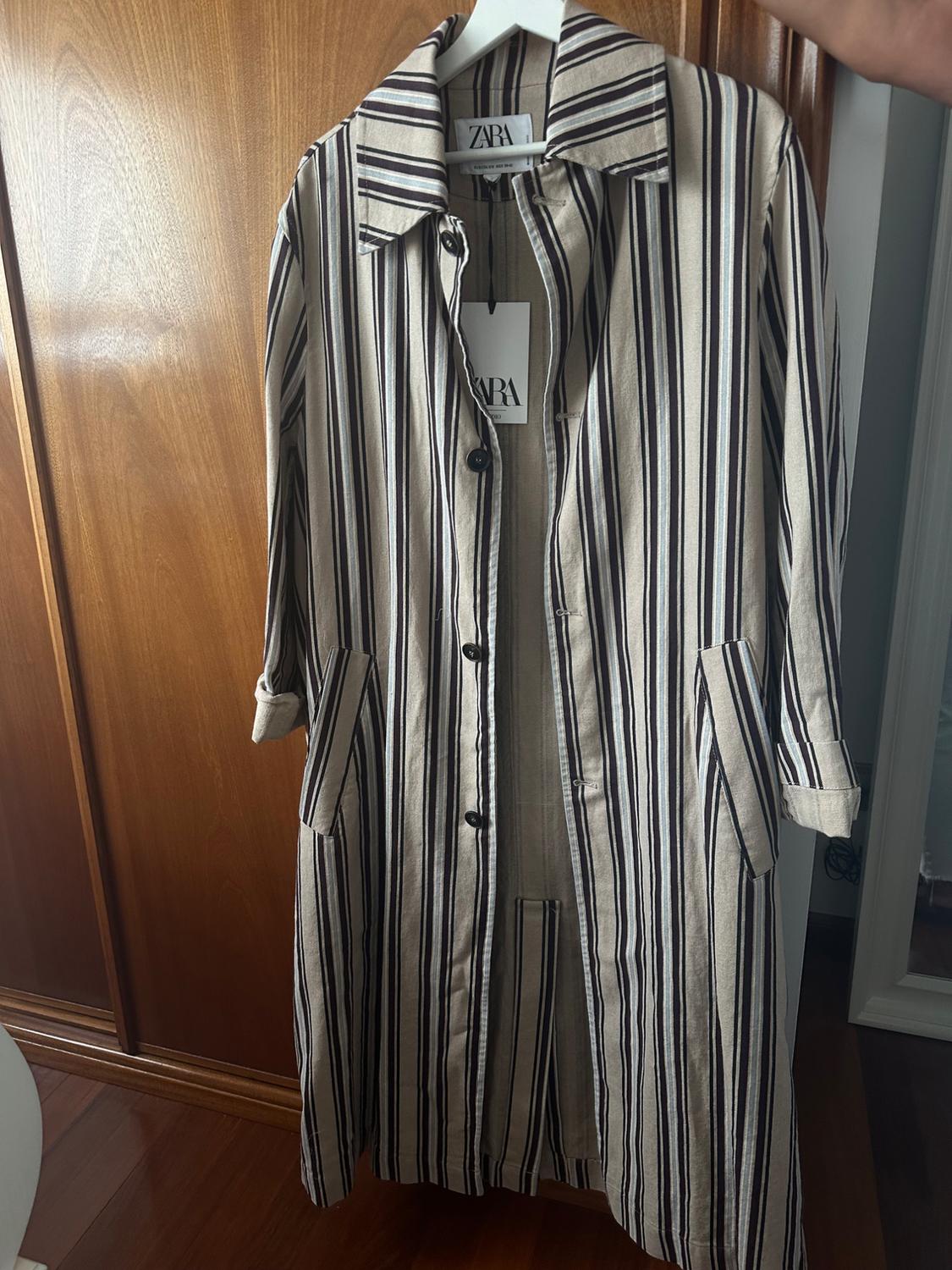 Vertical striped coat in beige and black tones hanging on a white hanger. Visible label with the text "ZARA".