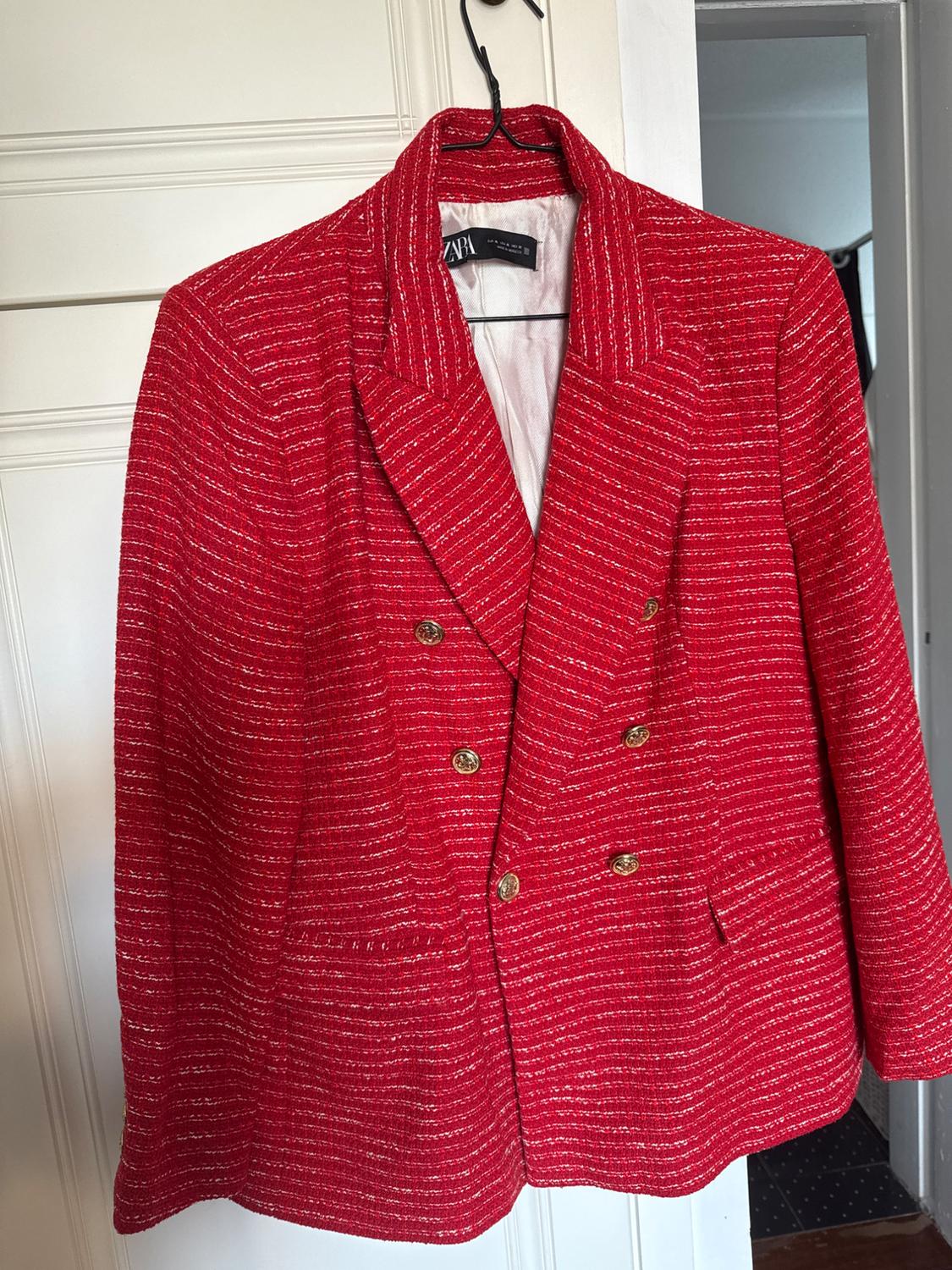 Red jacket with white stripes and gold buttons hanging on a black hanger. White wall background and a slightly ajar door.