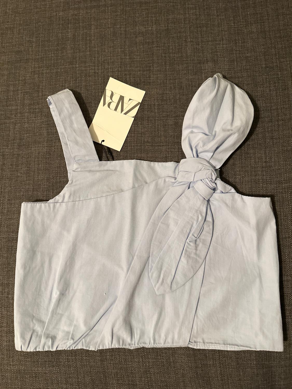 White top with one strap and decorative knot on a gray background. Label with "ZARA" text visible on the left side.