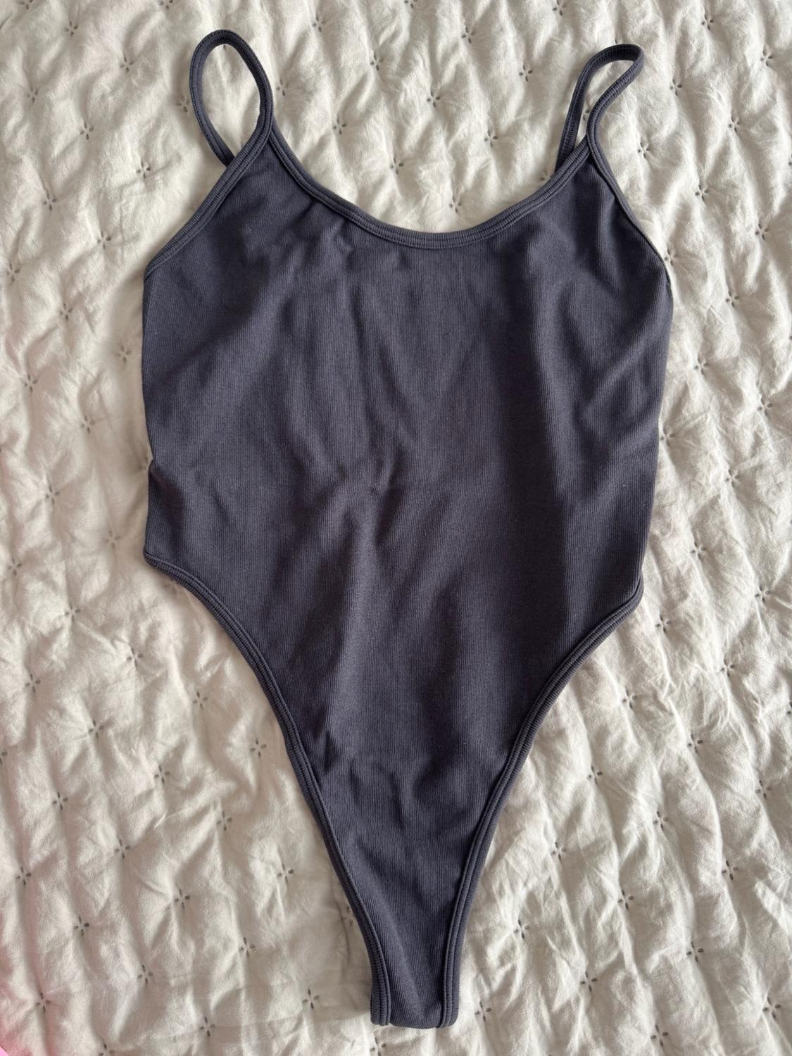 Dark grey ribbed bodysuit with thin straps, laid out on a textured white bedspread. Casual and relaxed style composition.