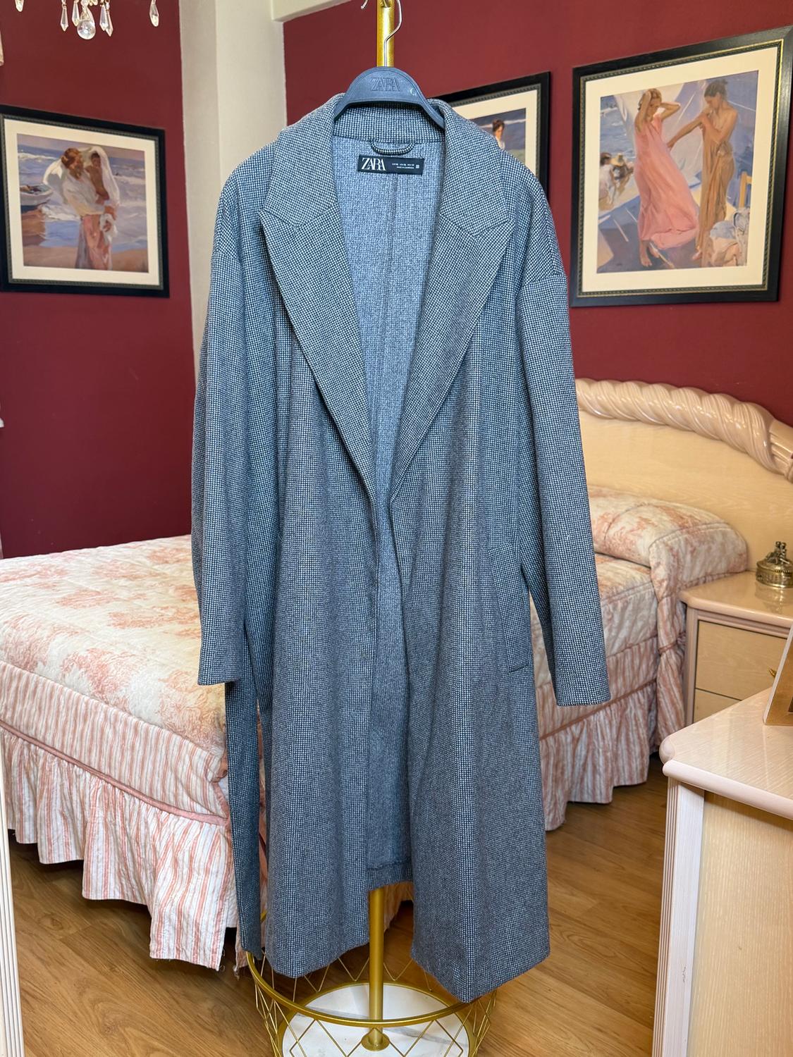 Grey coat hanging on a gold clothes rail in a bedroom with red walls and pictures. Visible label: "ZARA".