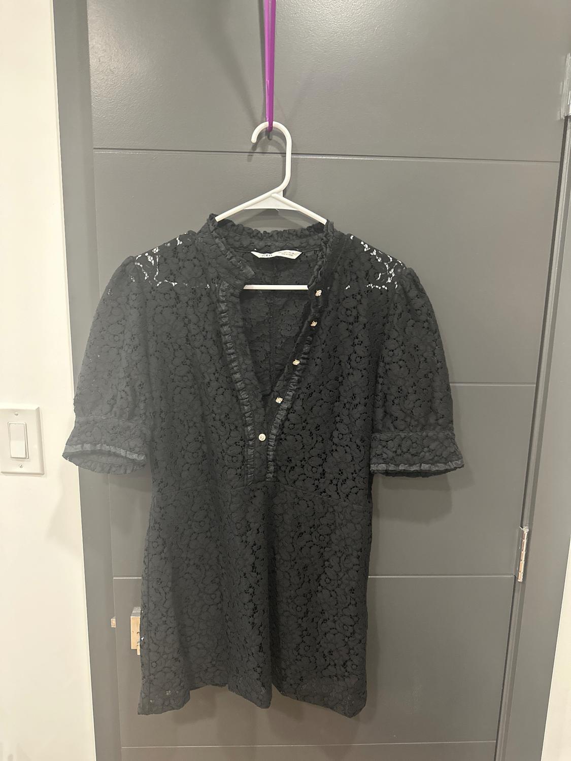 Black lace blouse hanging on a white hanger with a purple hook, over a gray door. Button detail on the front.