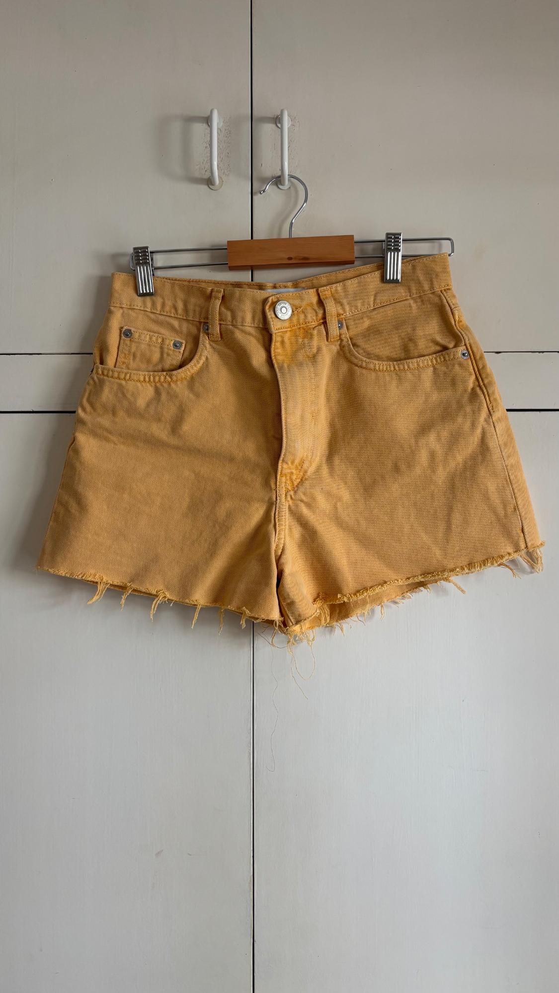 Yellow denim shorts with a frayed hem, hanging on a wooden hanger in front of a white wardrobe. Casual and relaxed style.