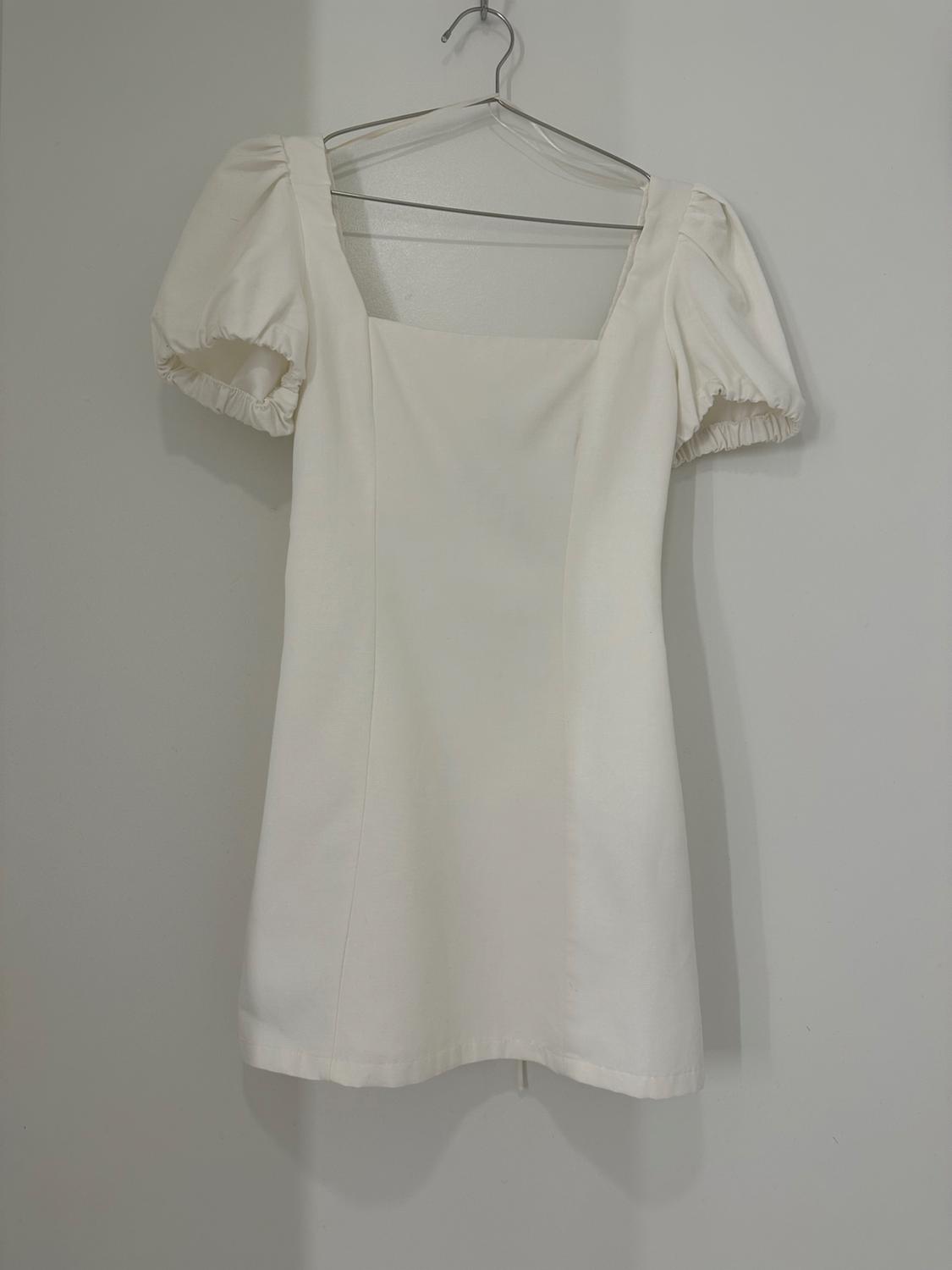 White short dress with a square neckline and short puff sleeves, hanging on a metal hanger in front of a plain white wall.