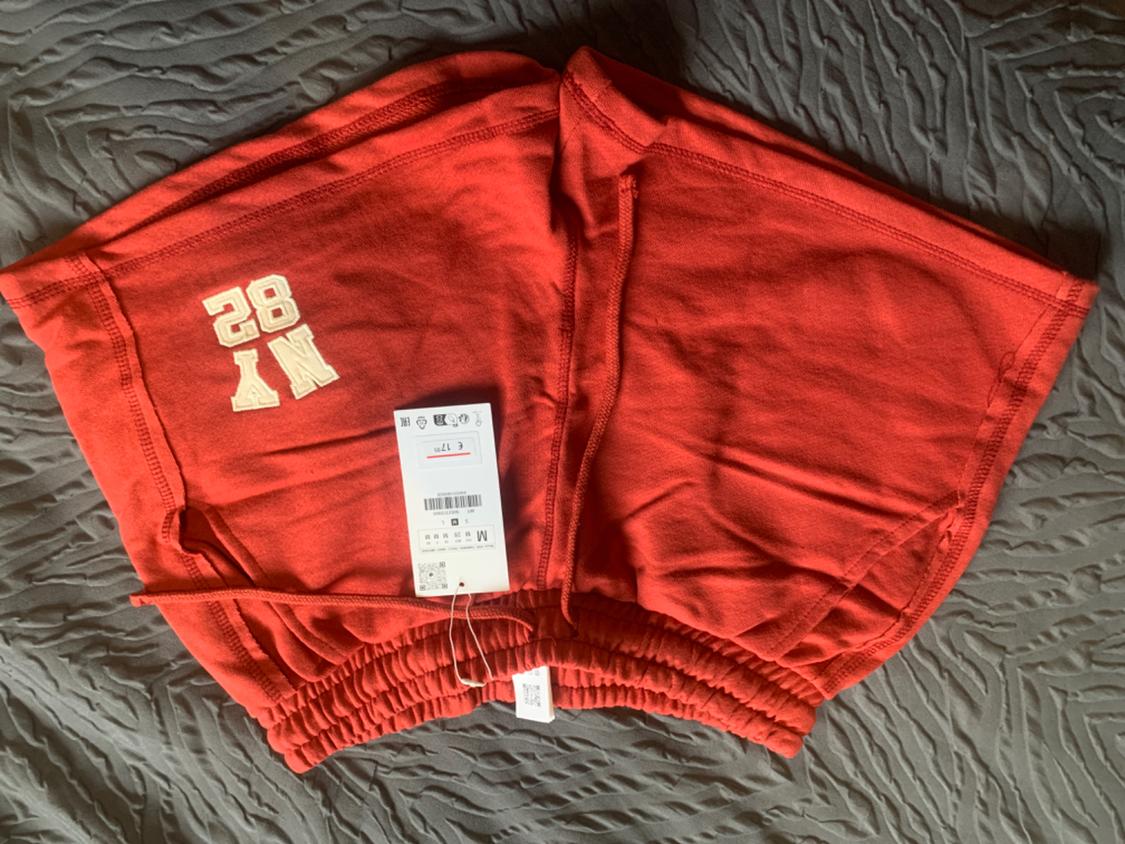 Red shorts with the number 82 and white NY lettering. Visible label with size and price details on a grey surface.