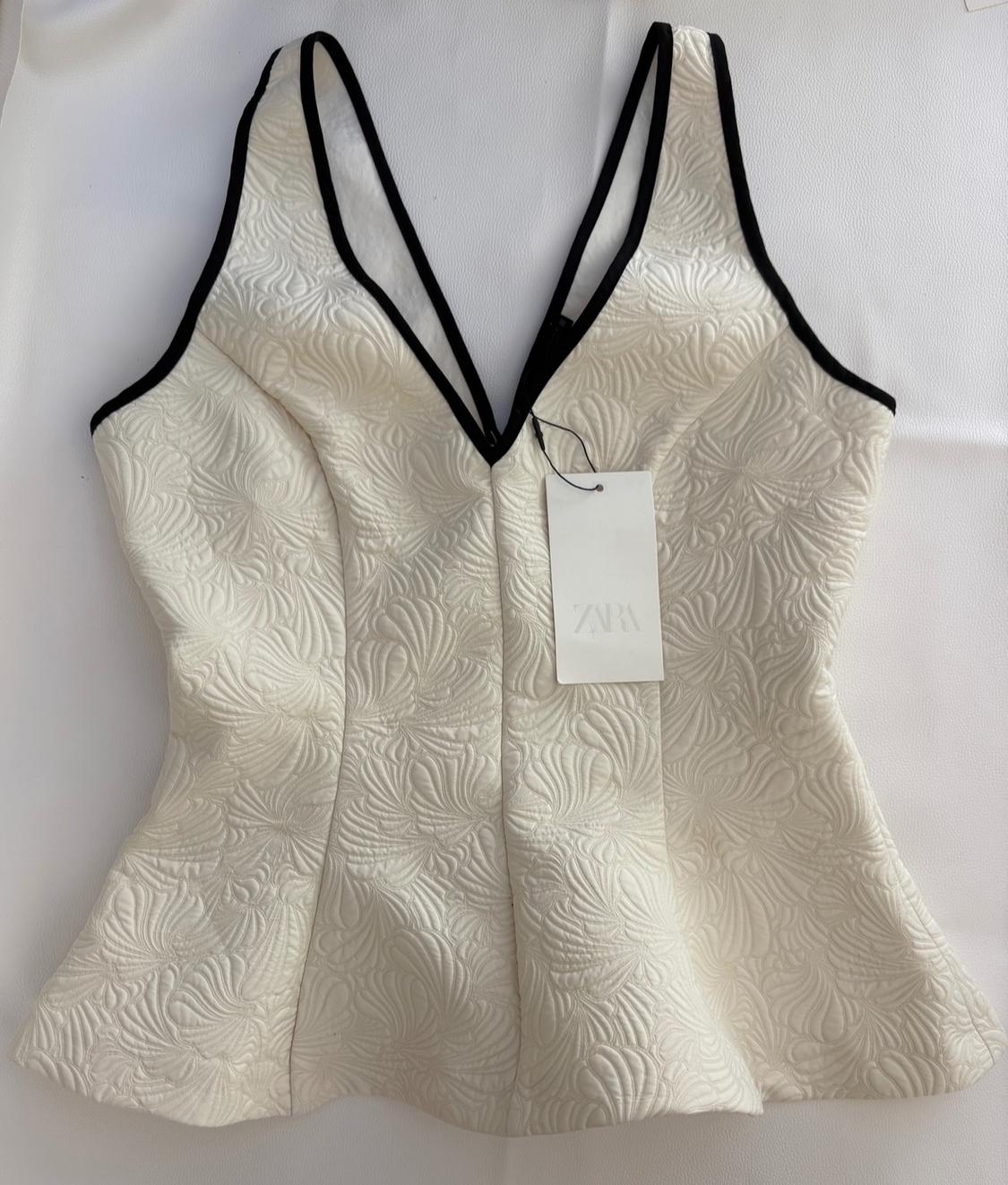 Textured white top with a V-neck and black trim. Flared hem. Label with the text ZARA. Presented on a white background.
