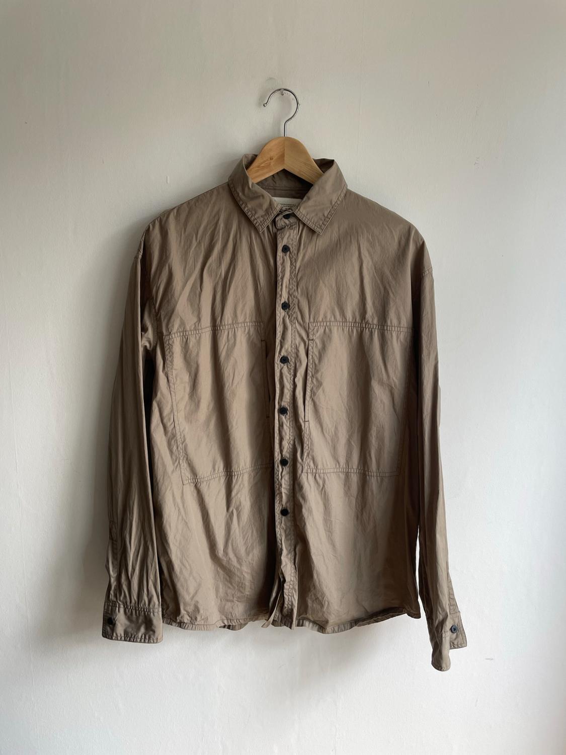 Long-sleeved beige shirt with dark buttons and front pockets, hanging on a wooden hanger against a white wall.