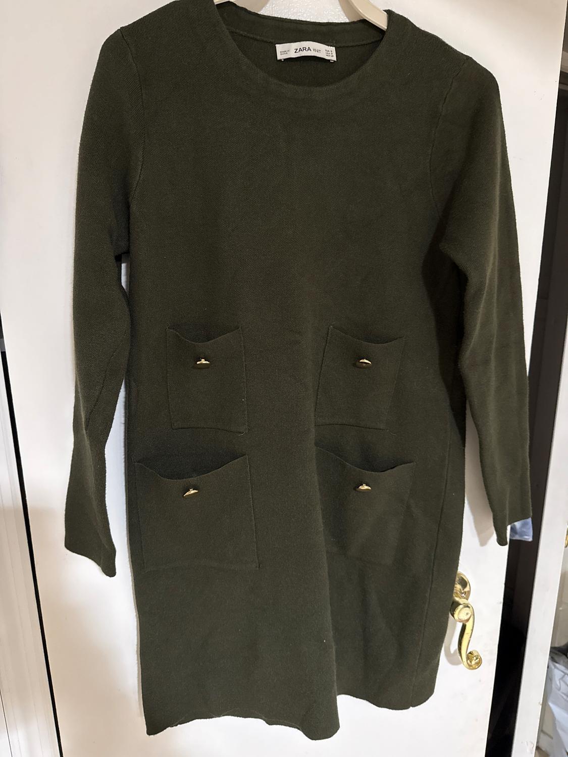Olive green long-sleeved dress with four front pockets and gold buttons, hanging on a white door. Visible tag: ZARA.