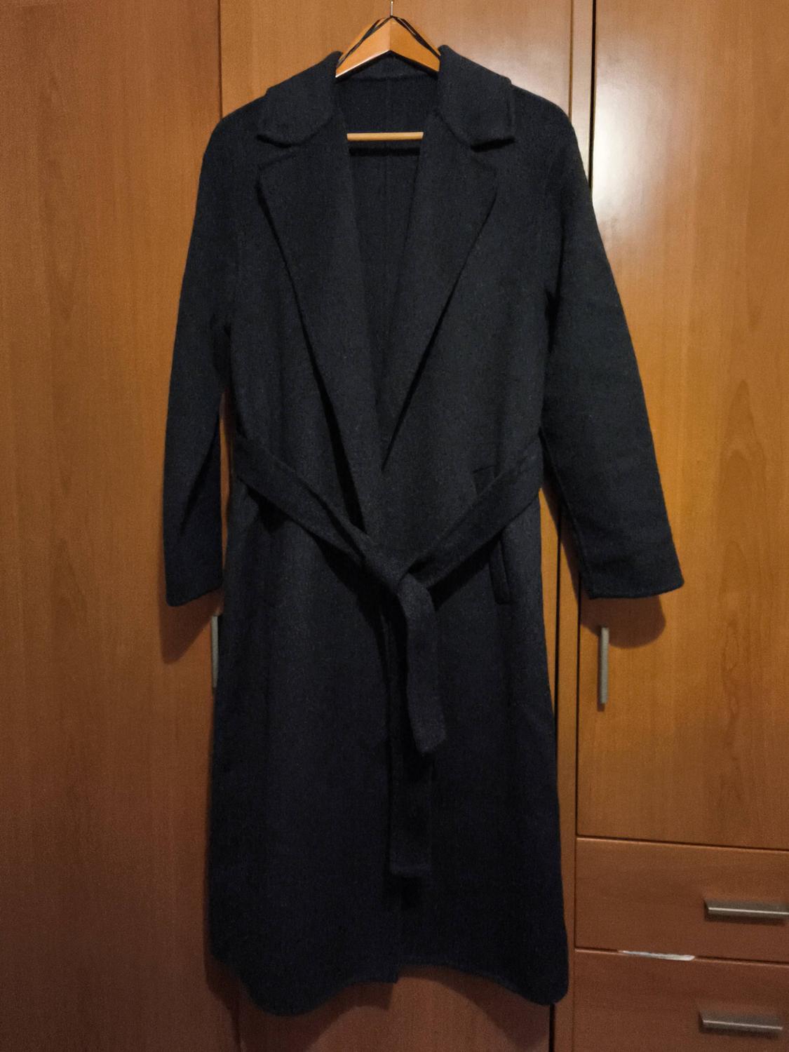 Long black coat with a belt, hanging on a wooden hanger, in front of a light wood wardrobe.