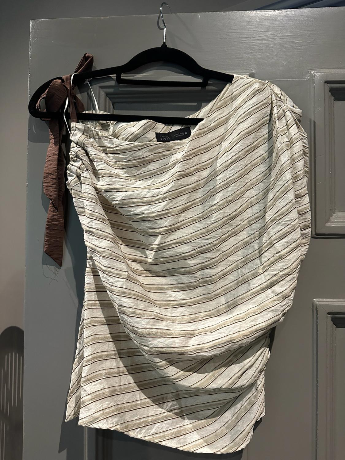 Draped striped top with a brown bow hanging on a hanger on a grey door. Text ZARA.