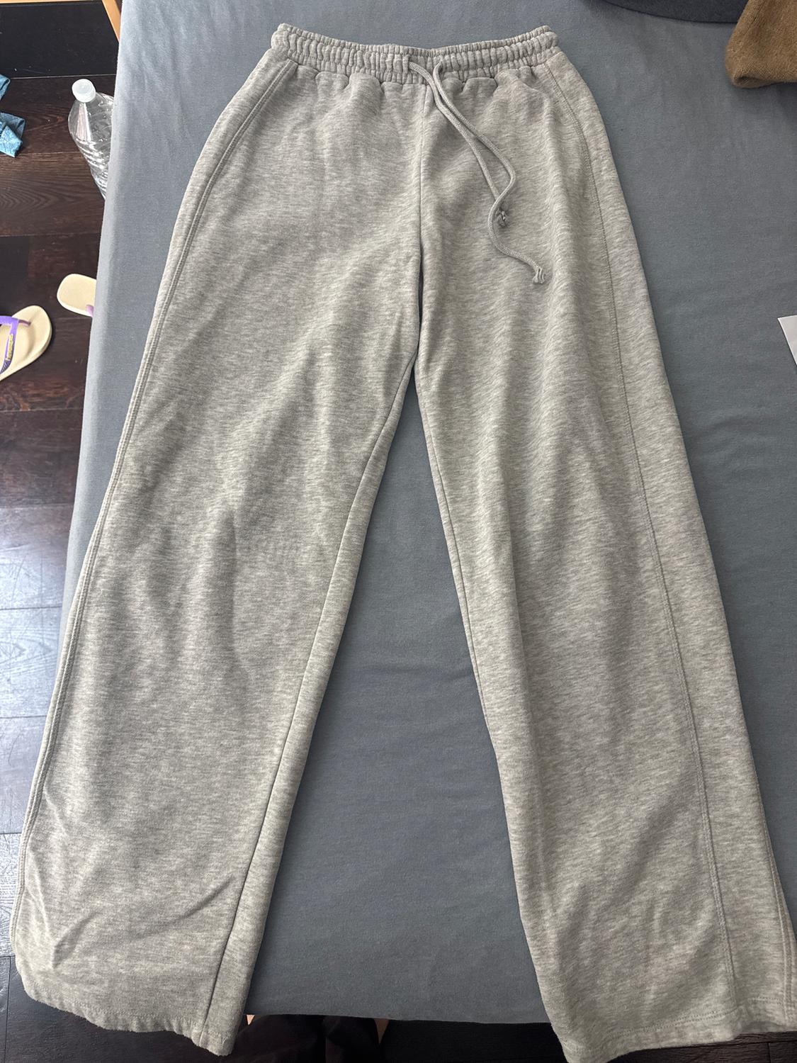 Grey jogging bottoms with an elasticated waist and drawstring, placed on a grey bed. Water bottle and sandals in the background.