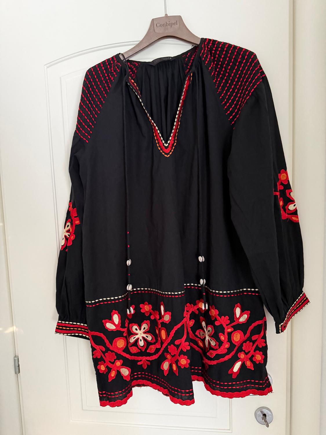 Black blouse with red and white embroidery, hanging on a wooden hanger with the "Conbipel" brand visible.