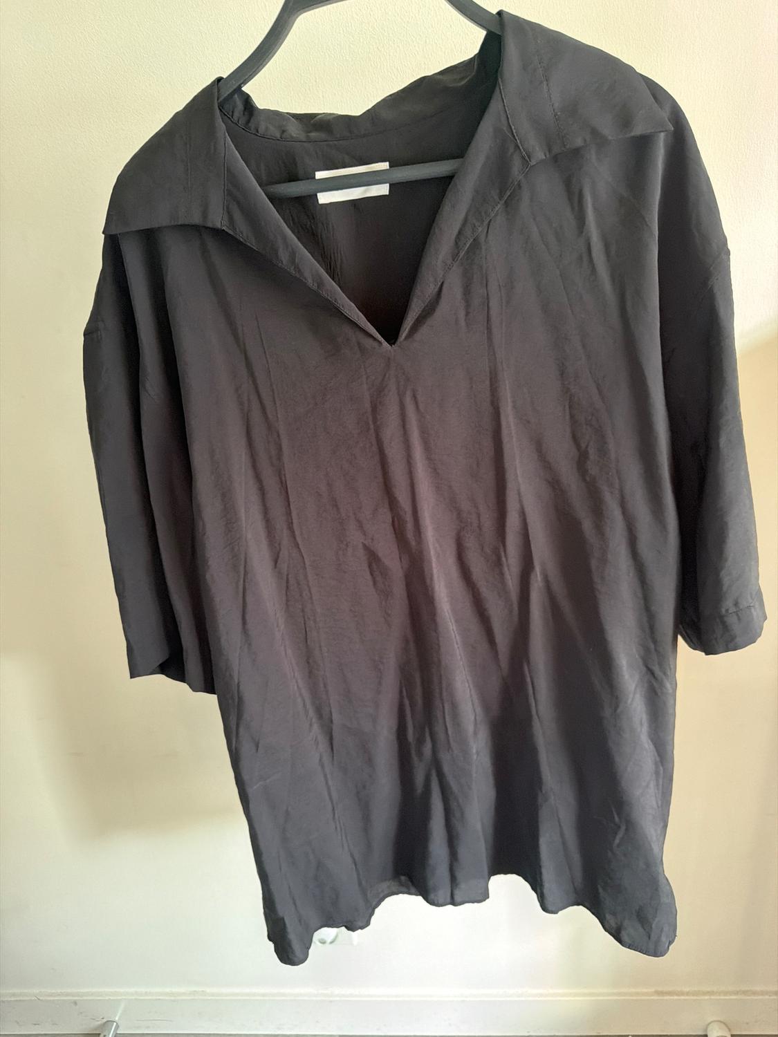 Black long-sleeved shirt hanging on a hanger, with an open collar and wrinkled fabric, against a light background.