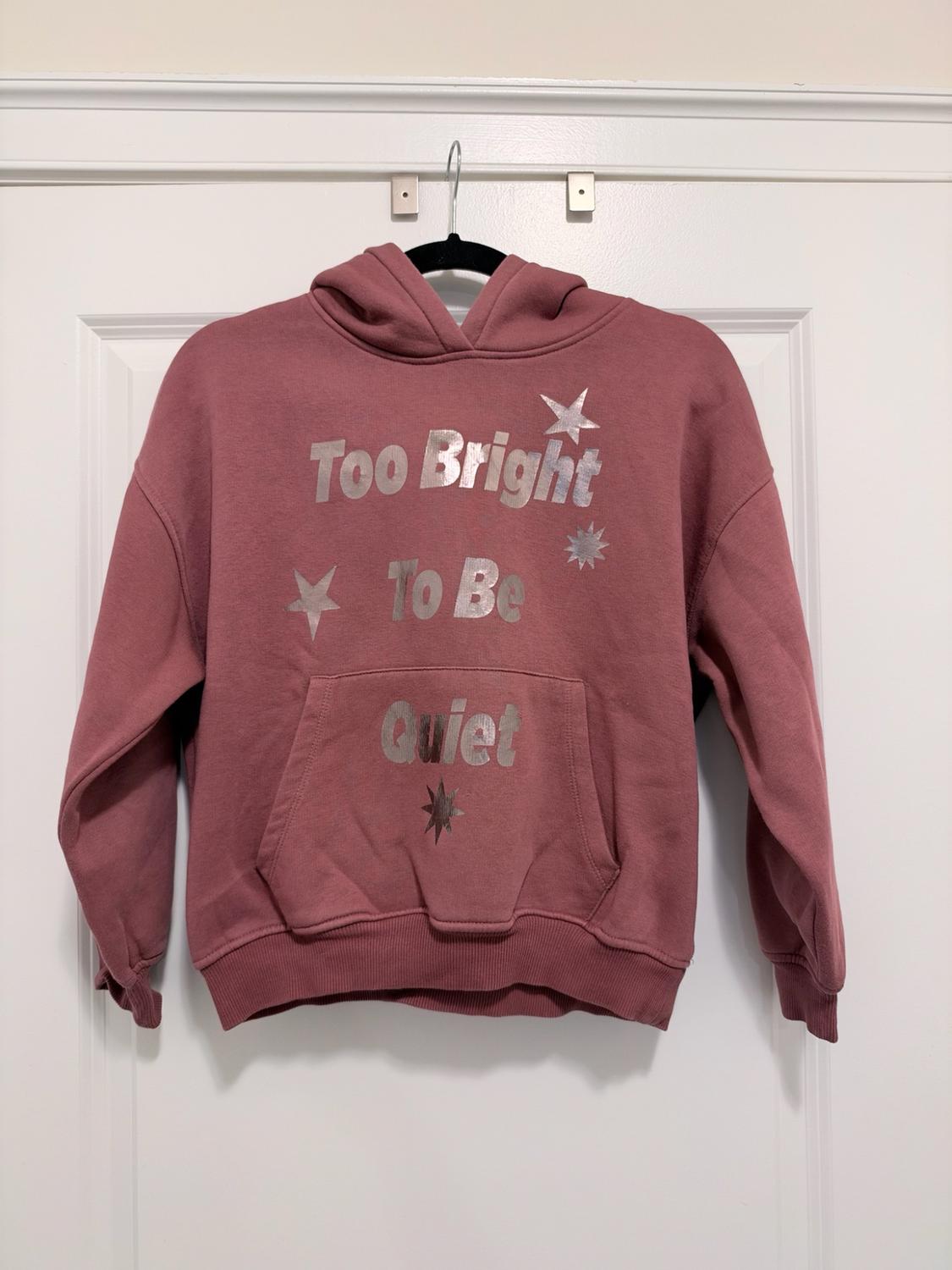 Pink hoodie with a pocket hanging on a white door. It features the silver text "Too Bright To Be Quiet" and stars.