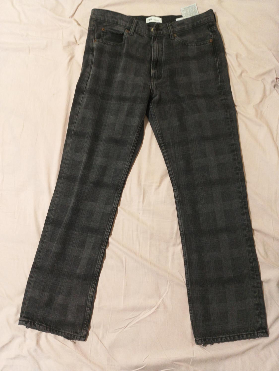 Dark denim jeans with a subtle checked pattern on a light surface. Visible label at the top.