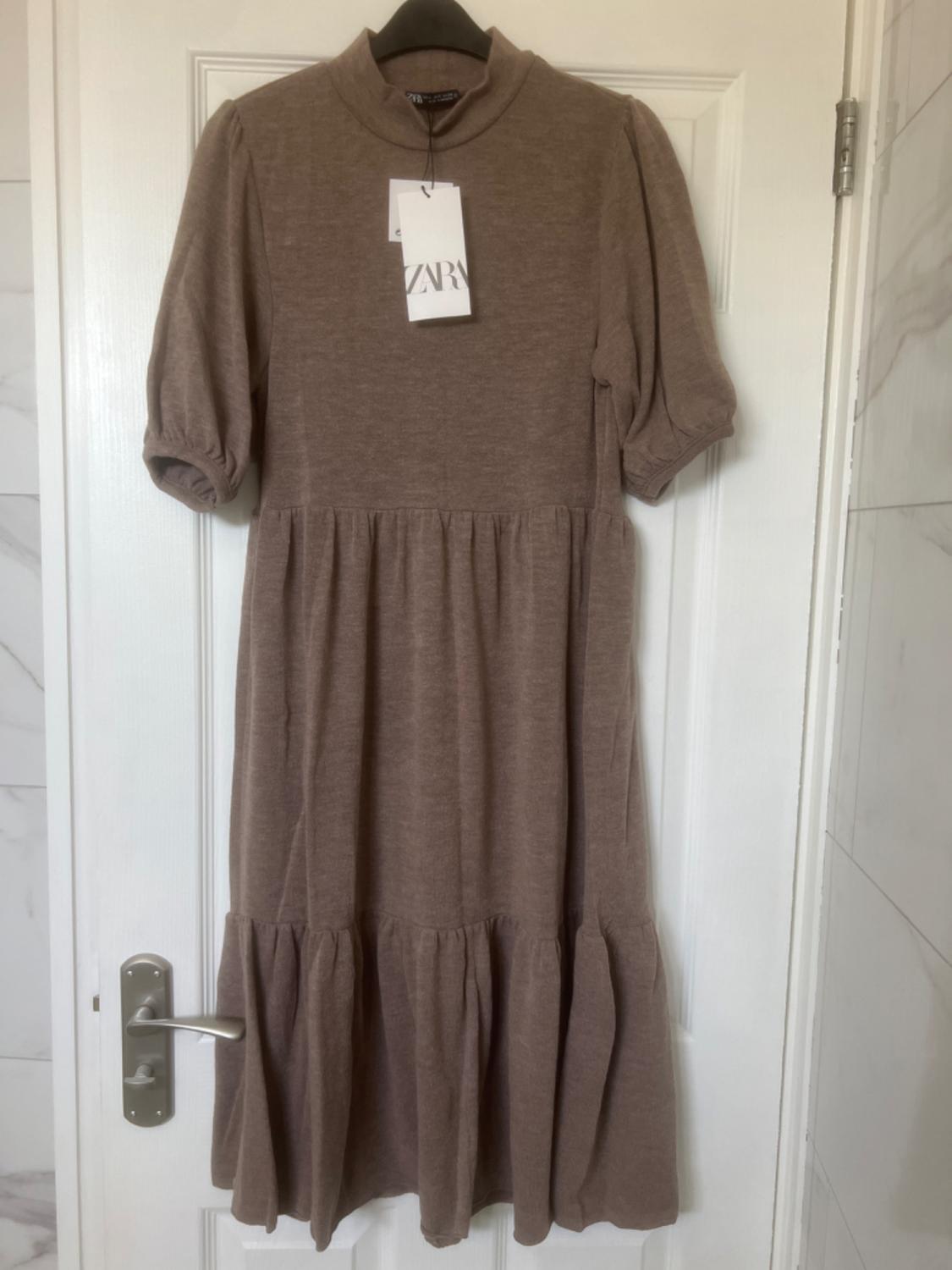 Brown short-sleeved dress with a high neck, hanging on a white door. Label visible with the text "ZARA".