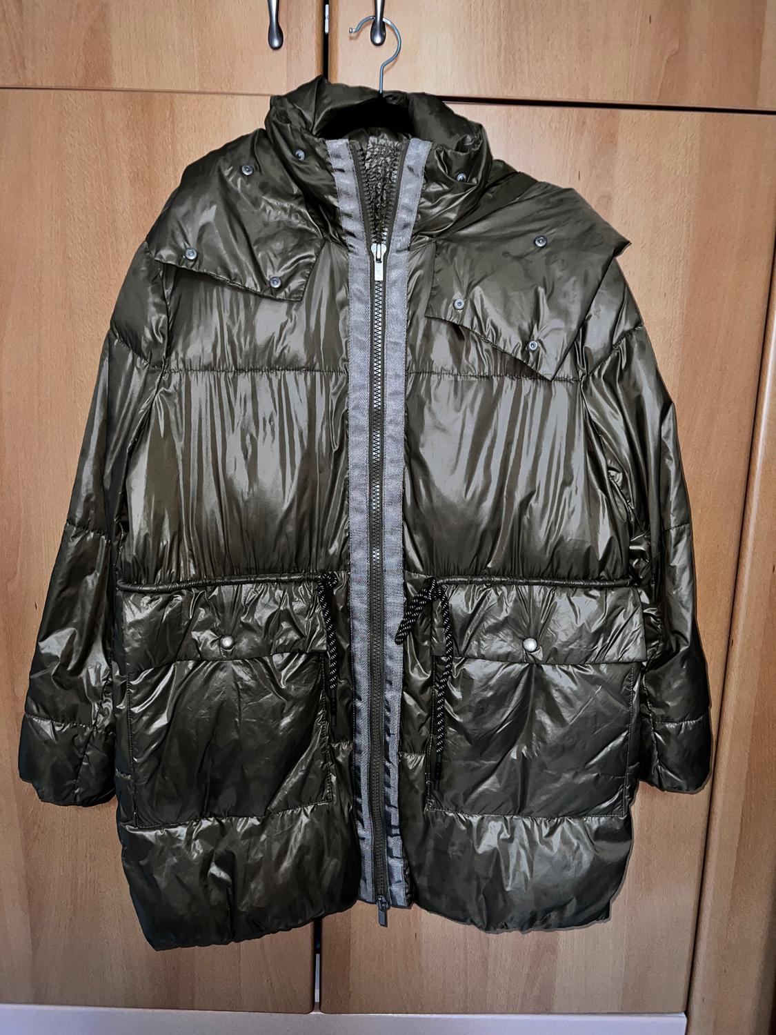 Olive green padded jacket hanging on a coat hanger, with a central zip and large pockets, against a light wood background.