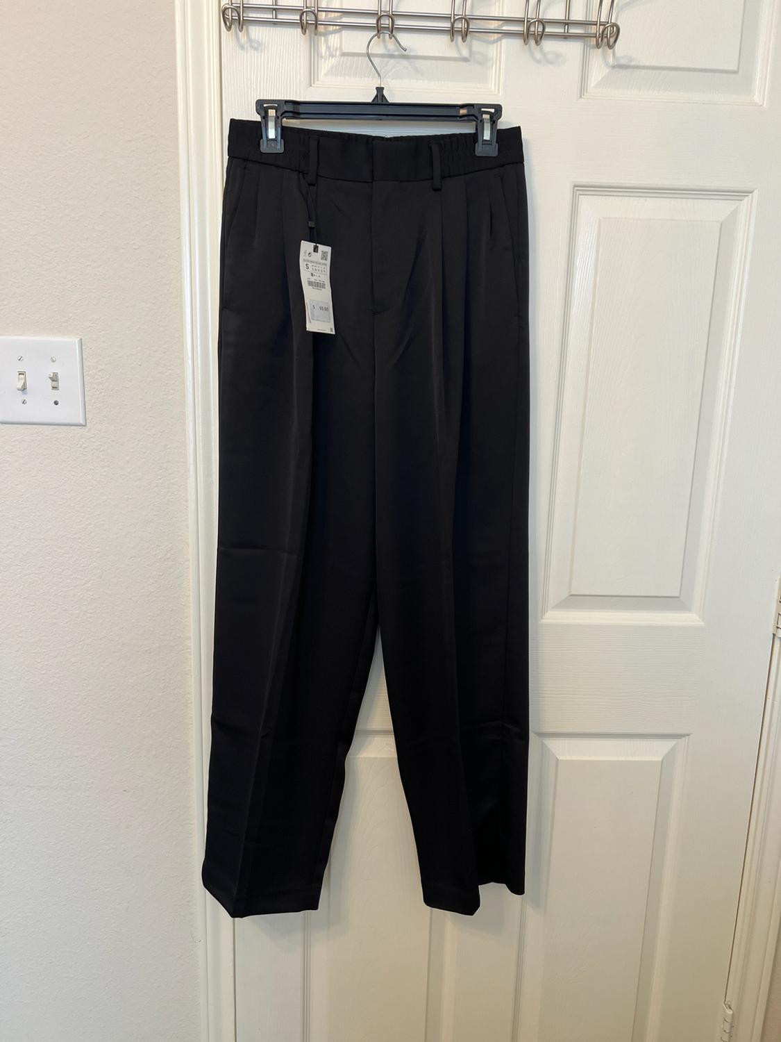 Black pleated trousers hanging on a coat hanger over a white door. Label visible at the waist.
