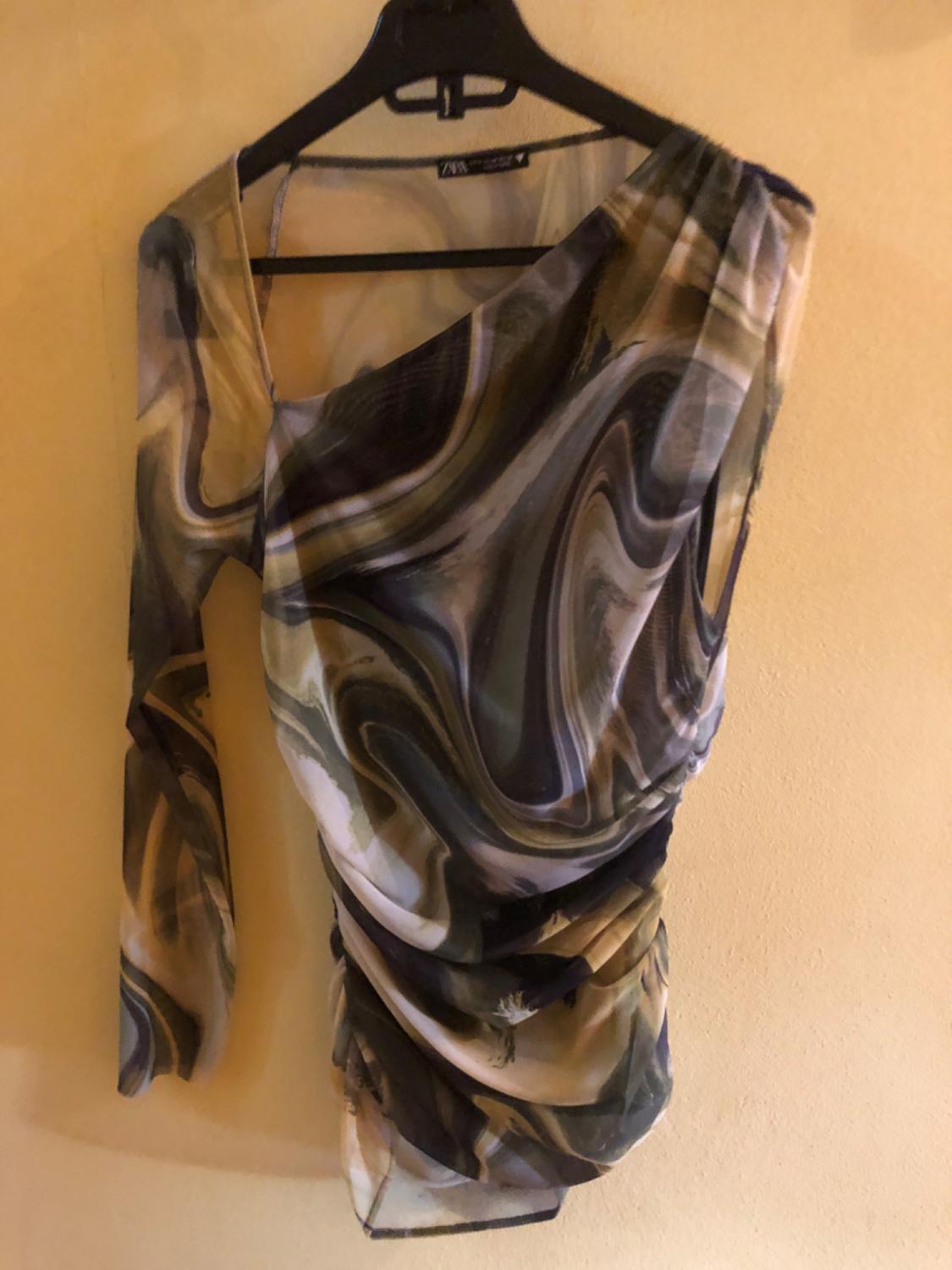 One-shoulder blouse with abstract print in brown and beige tones, hanging on a black hanger against a beige background.