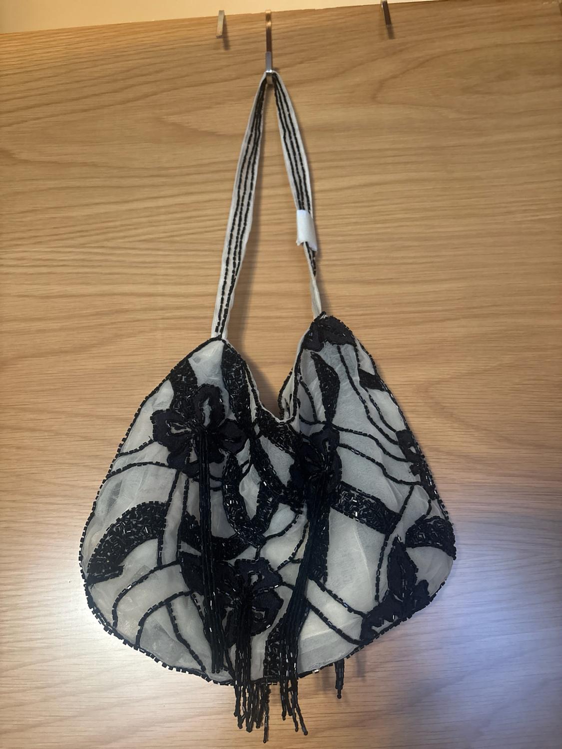 White handbag with floral details and black beaded fringing, hanging from a metal hook against a light wood background.
