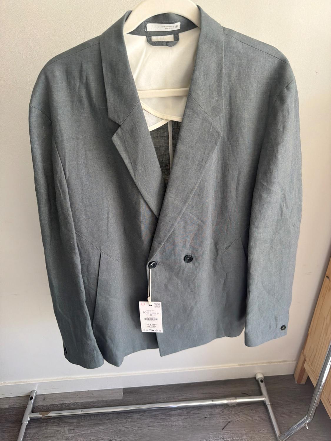 Grey linen jacket hanging on a white hanger, with a visible tag. White wall and wooden floor background.