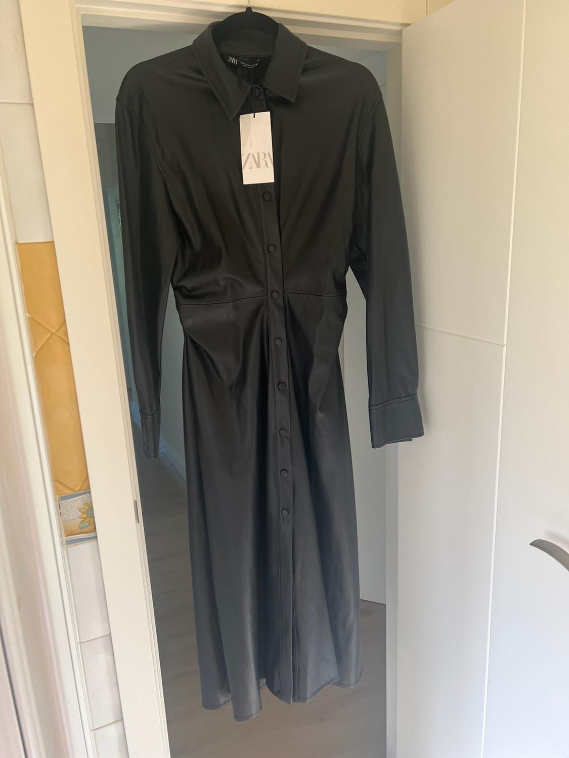 Long-sleeved black shirt dress hanging on a white door indoors. Includes a label bearing the text ZARA.